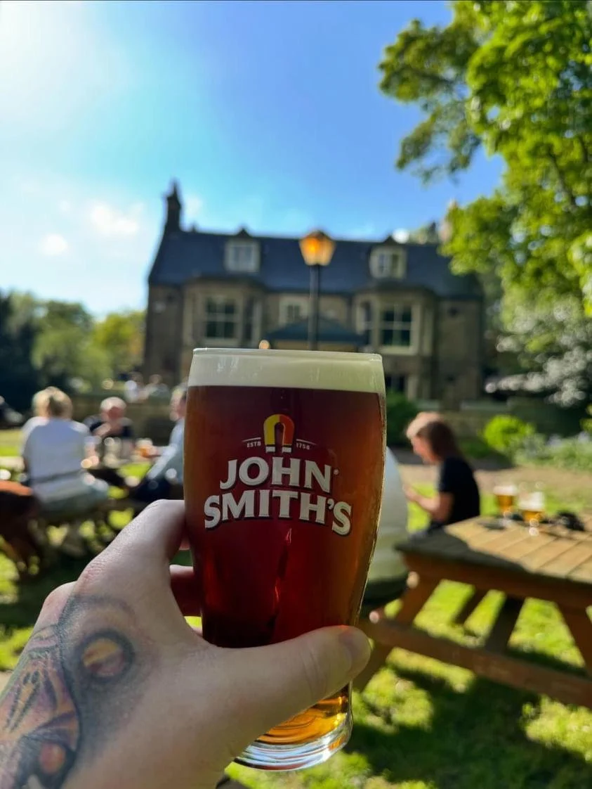 A hand holding a glass of John Smith's beer in an outdoor setting with a house, trees, and people in the background.