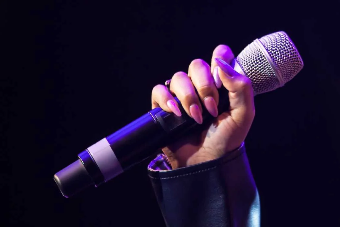 A hand with long pink nails holding a microphone against a dark background with purple lighting.