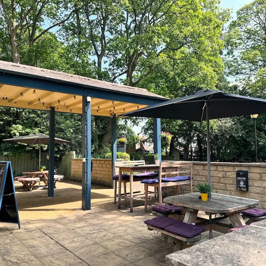 An outdoor patio with picnic tables, a large umbrella, hanging flower baskets, and lush green trees in the background.