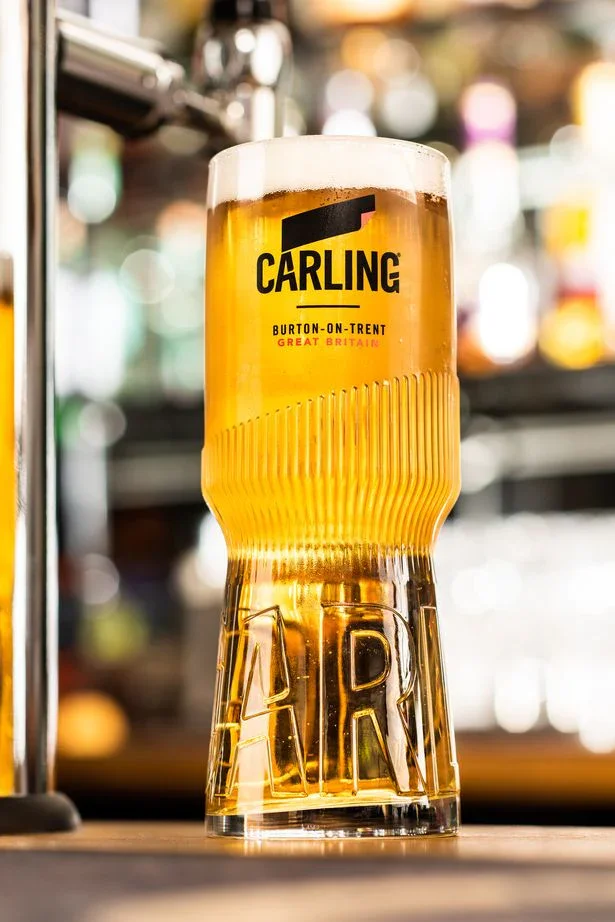 A glass of Carling beer with a layer of foam on top, placed on a bar counter with a blurred background of colorful lights.