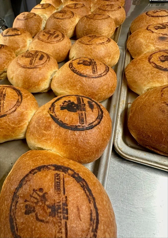 Baked bread rolls with stamped logos on top, arranged on metal trays.