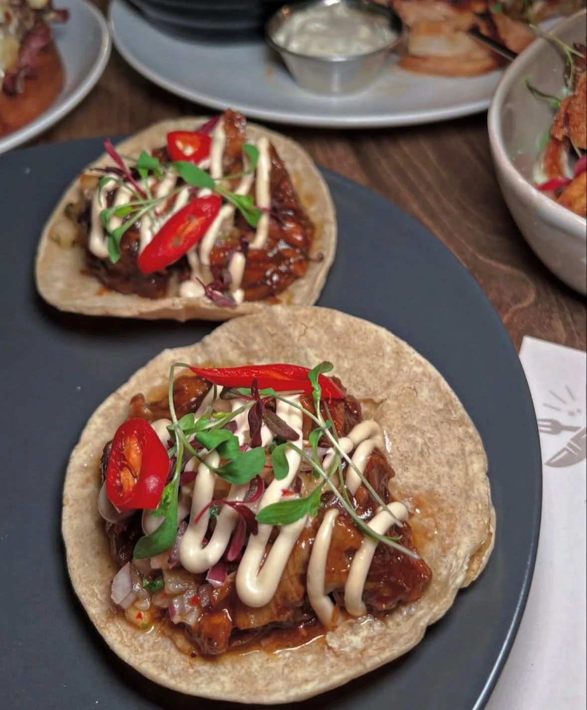 Two tacos on a dark plate topped with chopped red peppers, microgreens, and drizzled with mayonnaise and barbecue sauce, served with a side of dipping sauce.