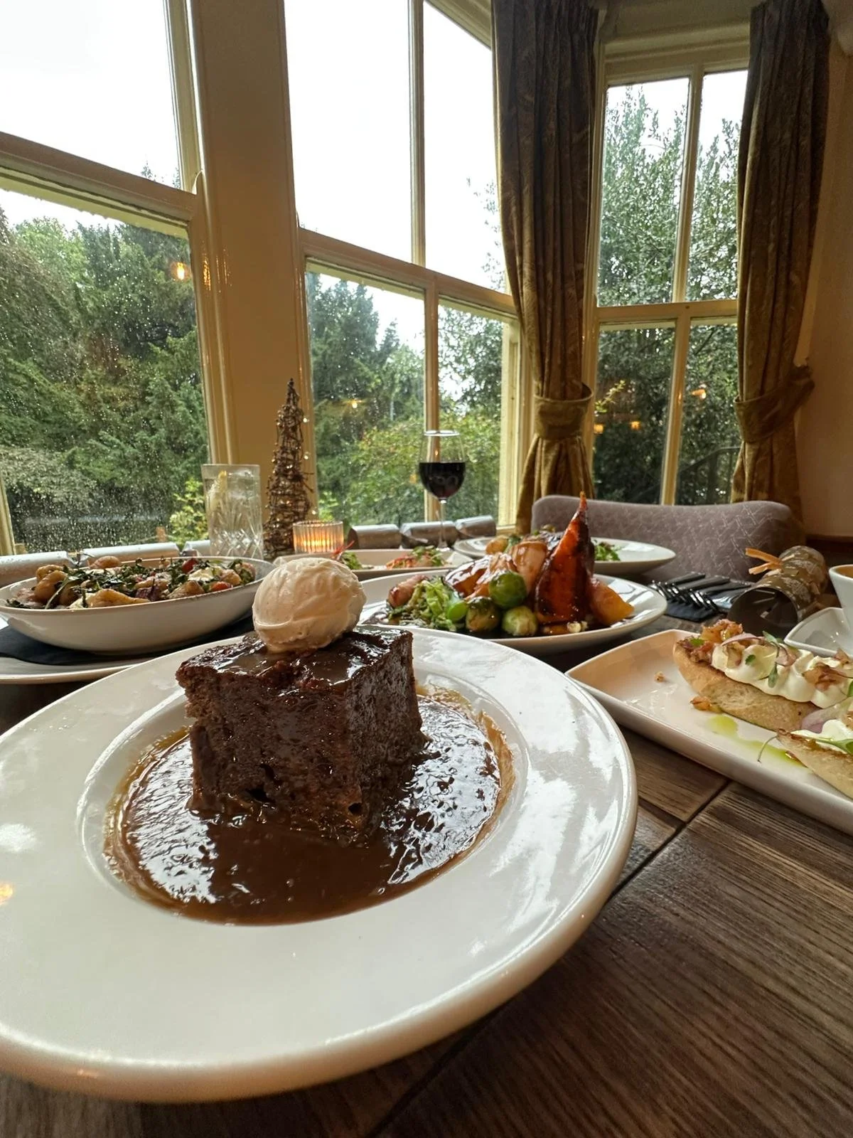 Close-up of a dessert plate with a chocolate cake square topped with a scoop of vanilla ice cream, surrounded by chocolate sauce, on a white plate. In the background, other dishes including a roasted chicken with vegetables, a salad, and another entrée are visible, set on a rustic wooden table with a window showing rain and green trees outside.