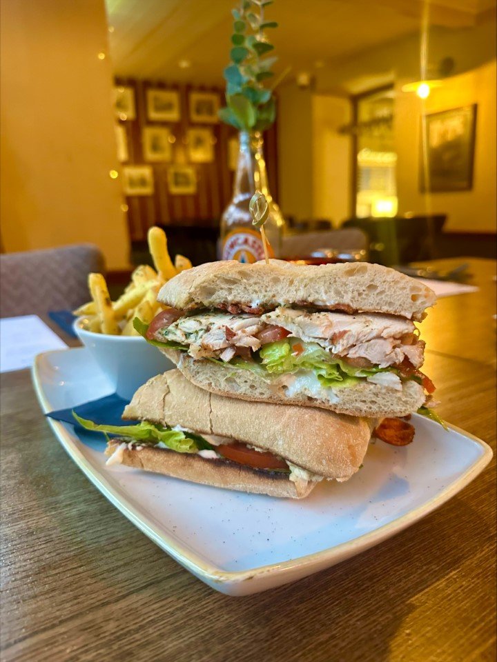 A club sandwich with lettuce, tomato, turkey, and bacon, served on a baguette, with a side of French fries in a small white cup on a white plate.