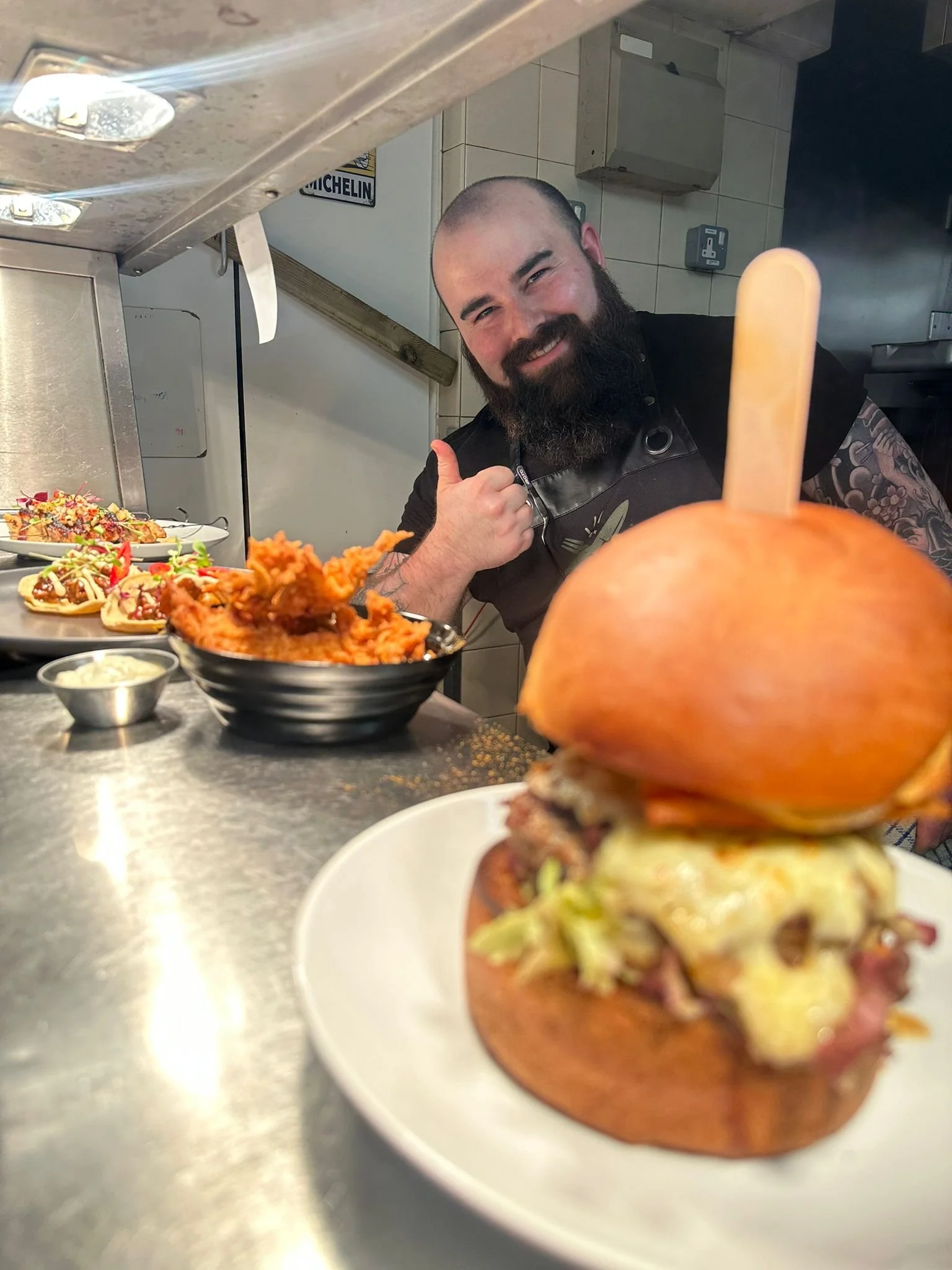 A chef smiling and giving a thumbs up behind a plate with a burger in a kitchen. The burger has lettuce, cheese, bacon, and a large bun with a popsicle stick on top.