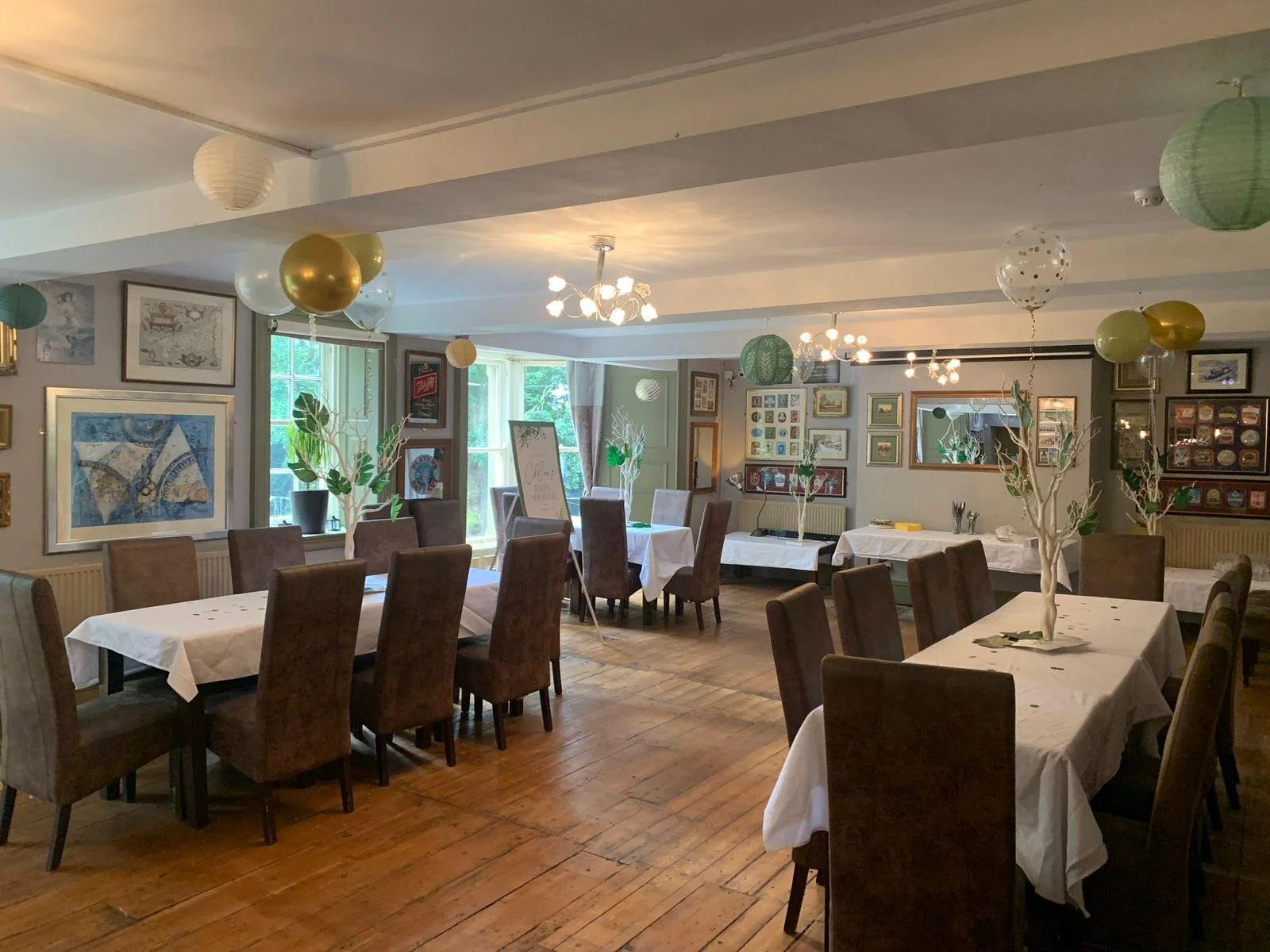 Decorated banquet hall with tables covered in white tablecloths, surrounded by brown chairs, featuring birthday balloons and festive decor.