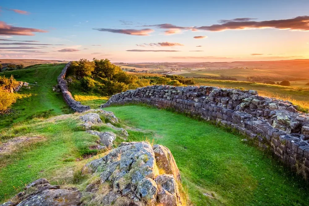 Sunset over ancient stone ruins with green grass and trees in the foreground and rolling hills in the distance.
