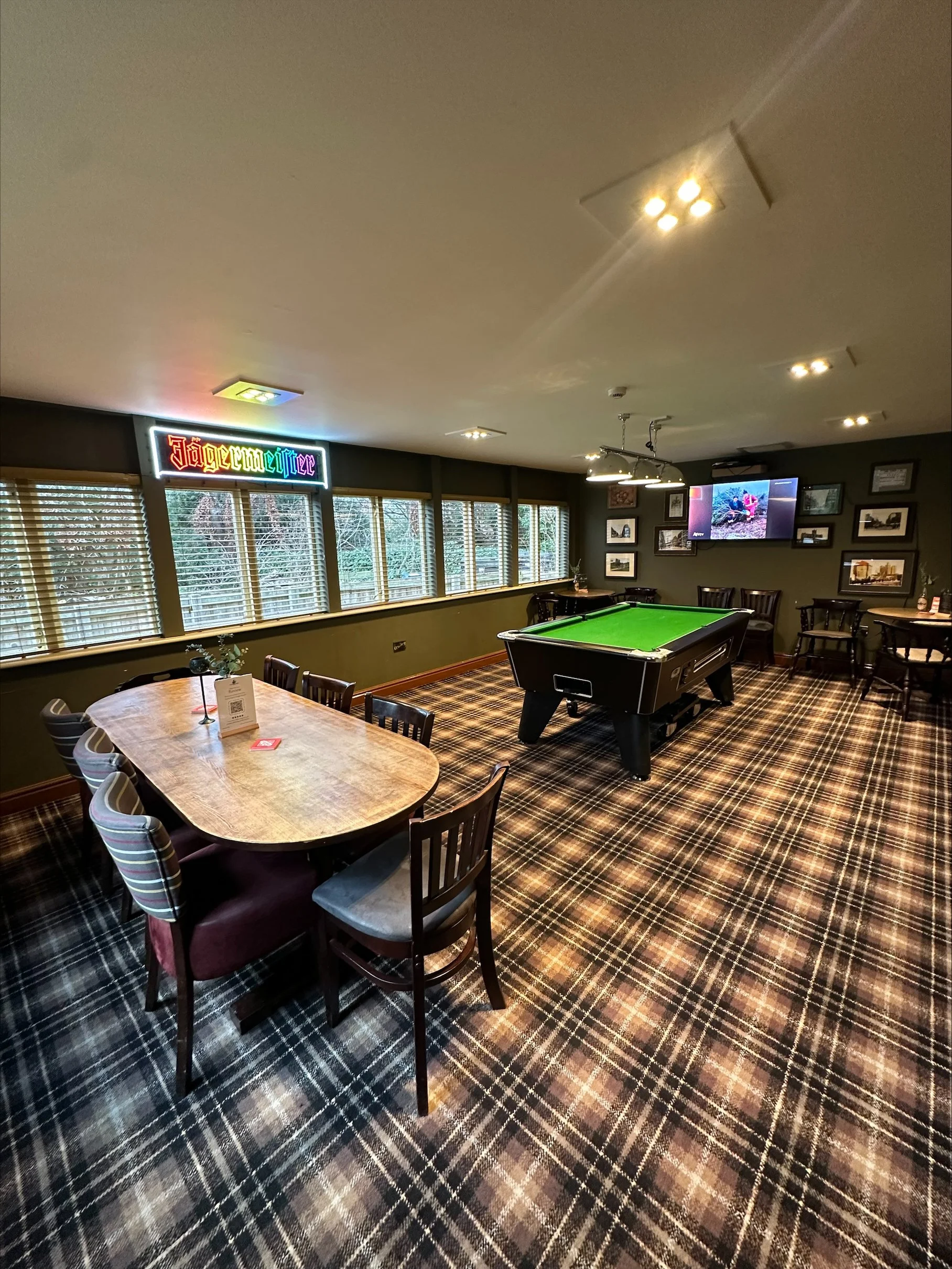 Interior of a game room with a pool table and a round dining table with chairs, decorated with framed pictures, a television, and a neon Jägermeister sign, with windows along one wall and plaid carpeting.