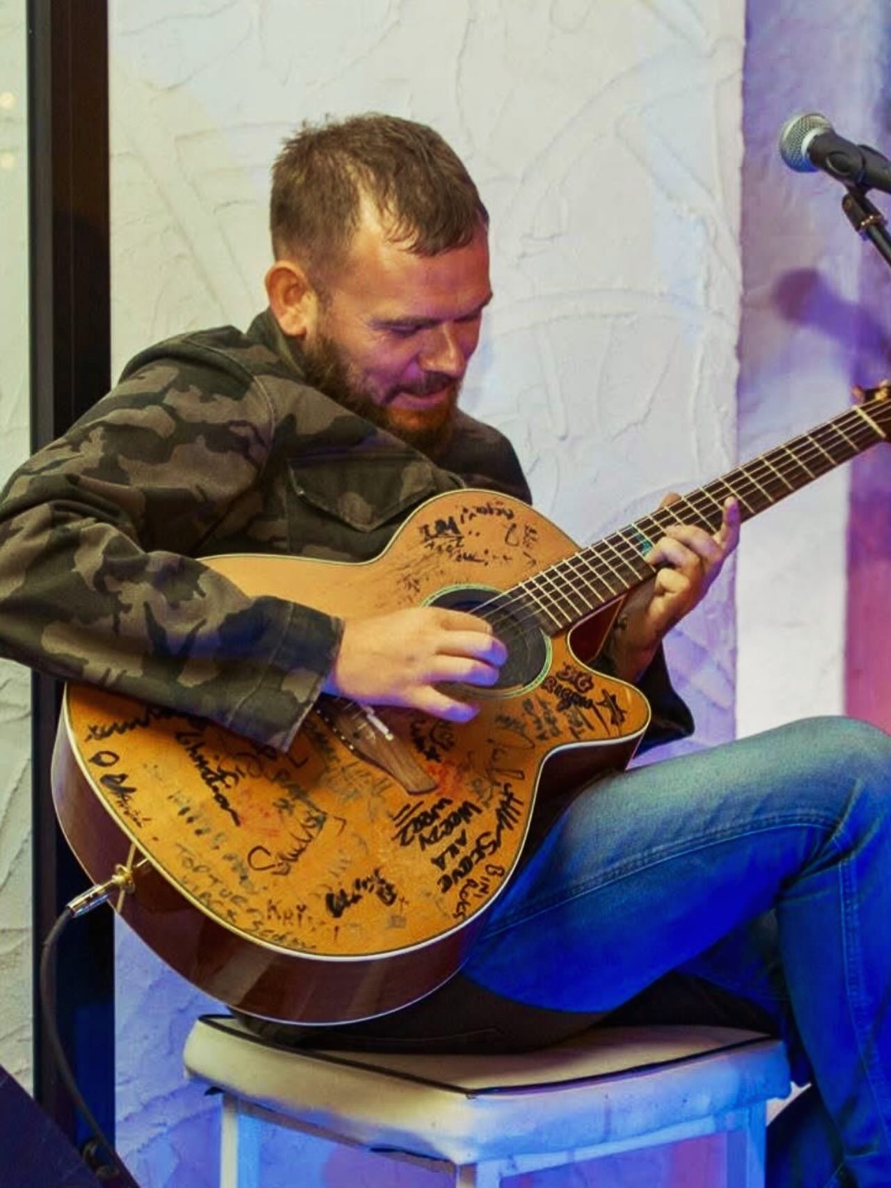 Man playing an acoustic guitar covered in signatures and messages, sitting on a stool with a microphone nearby.