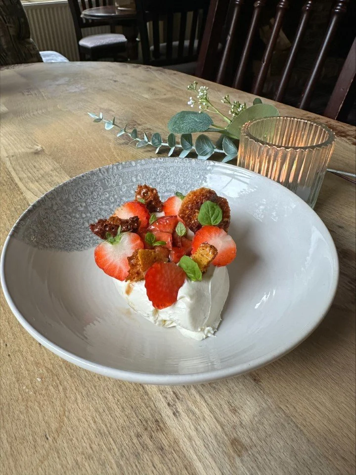 Dessert with strawberries, whipped cream, crumbles, and small green garnishes served on a white bowl, placed on a wooden table with a candle and some greenery in the background.
