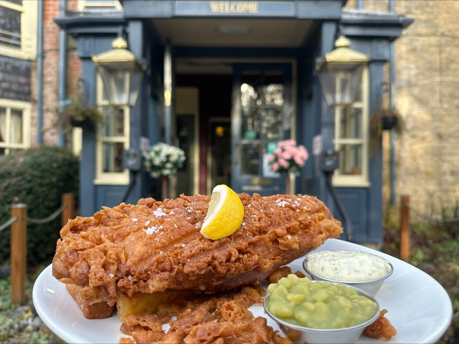 Plate of fried fish with lemon wedge, served with tartar sauce, green peas, and onion rings, in front of a blue building with lanterns and flowers.