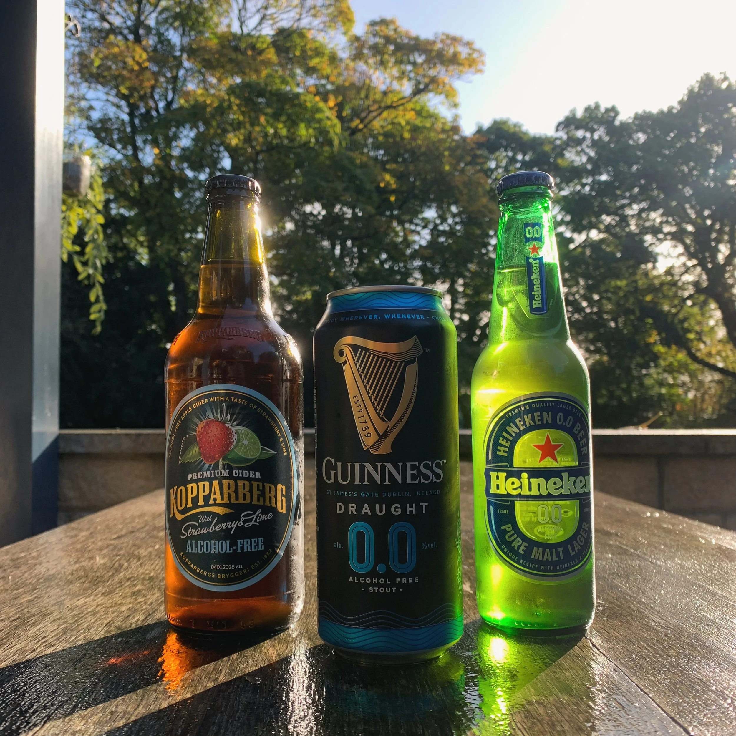 Three beverages on a wooden surface with a background of trees and sky. From left to right: a bottle of Koppaberg strawberry and lime cider, a can of Guinness alcohol-free stout, and a bottle of Heineken alcohol-free beer.