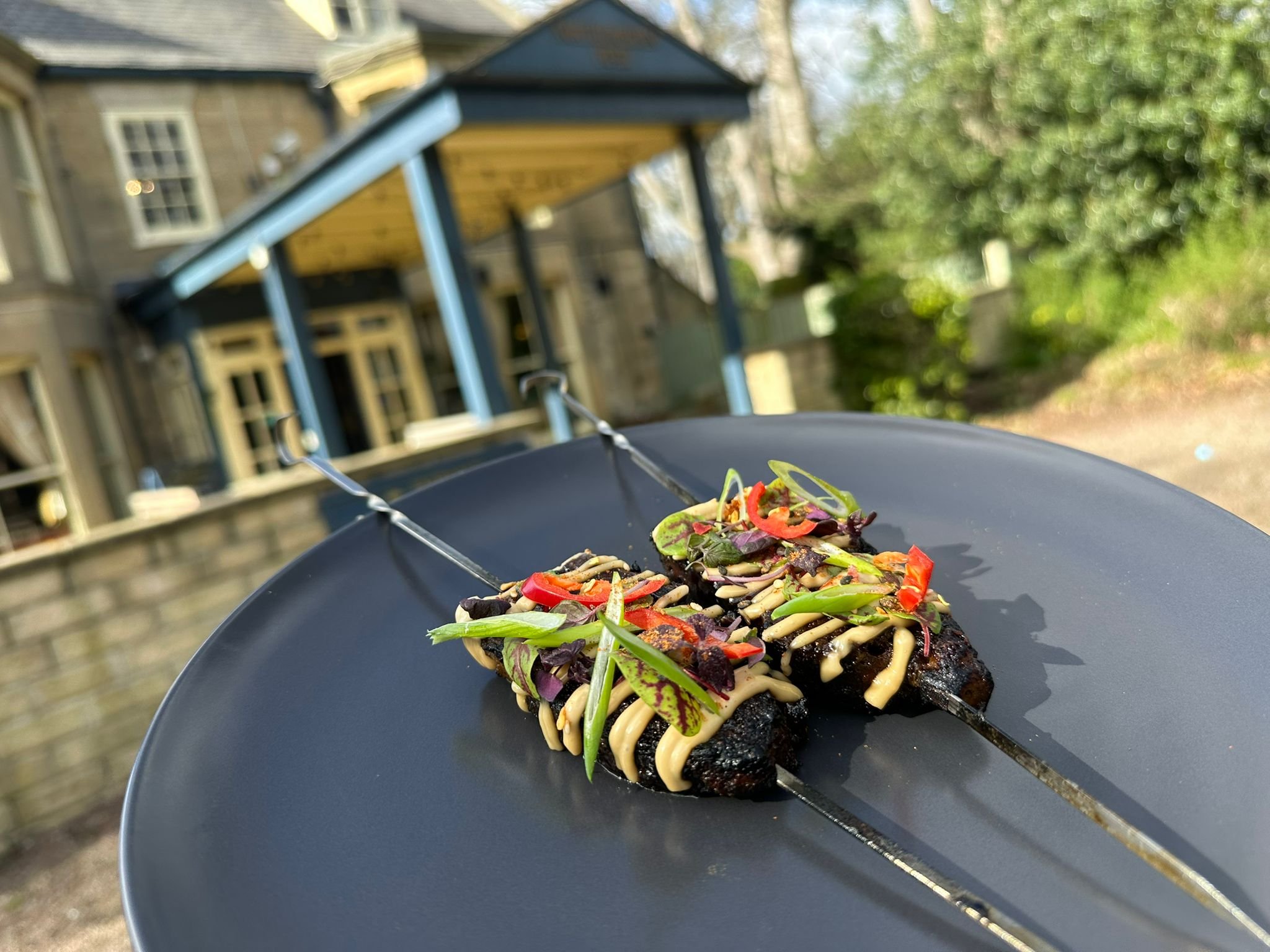 Two skewers of grilled meat topped with colorful vegetables, drizzled with sauce, on a black plate outdoors with a stone house and greenery in the background.