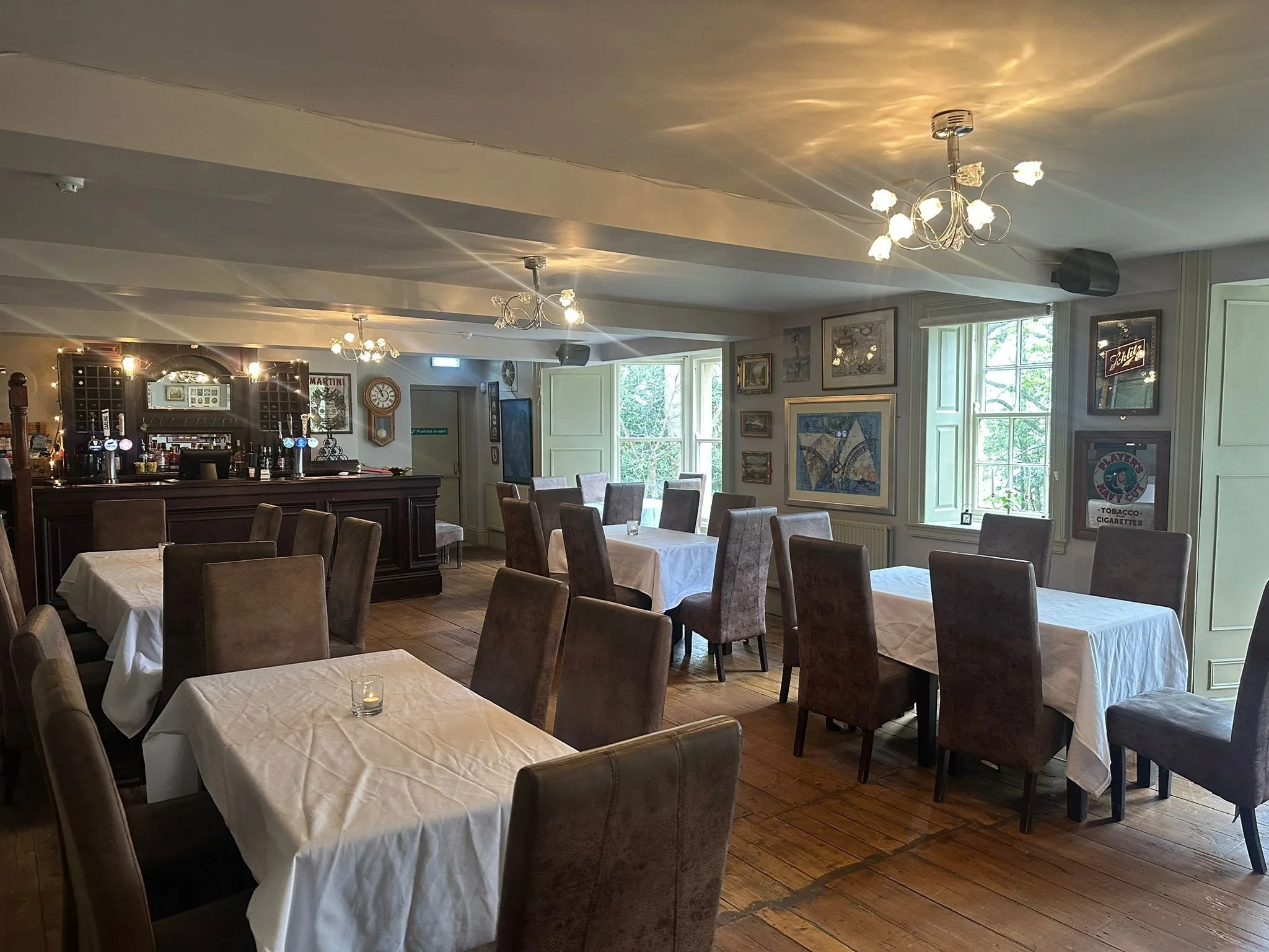A cozy restaurant with white tablecloth-covered tables, brown upholstered chairs, a bar area with bottles, wine rack, and framed artwork on the walls. Large windows let in natural light, and small decorative lights hang from the ceiling.