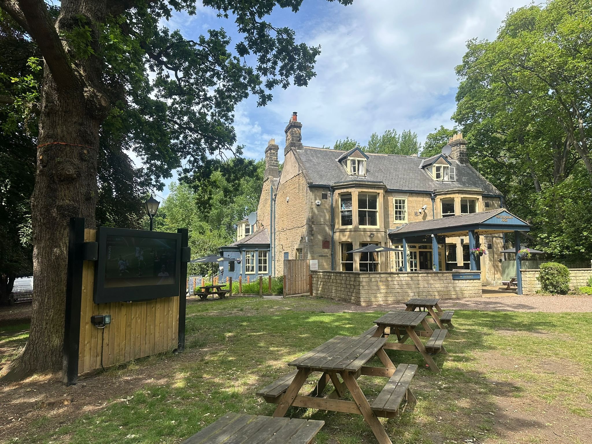A large, historic stone building with blue trim and multiple chimneys, surrounded by green trees, with an outdoor seating area with picnic tables in the foreground.