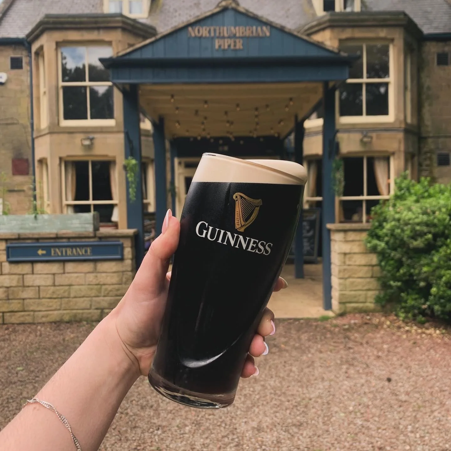 A person holding a tall glass of Guinness stout beer outside a building with a sign reading 'Northumbrian Piper'.