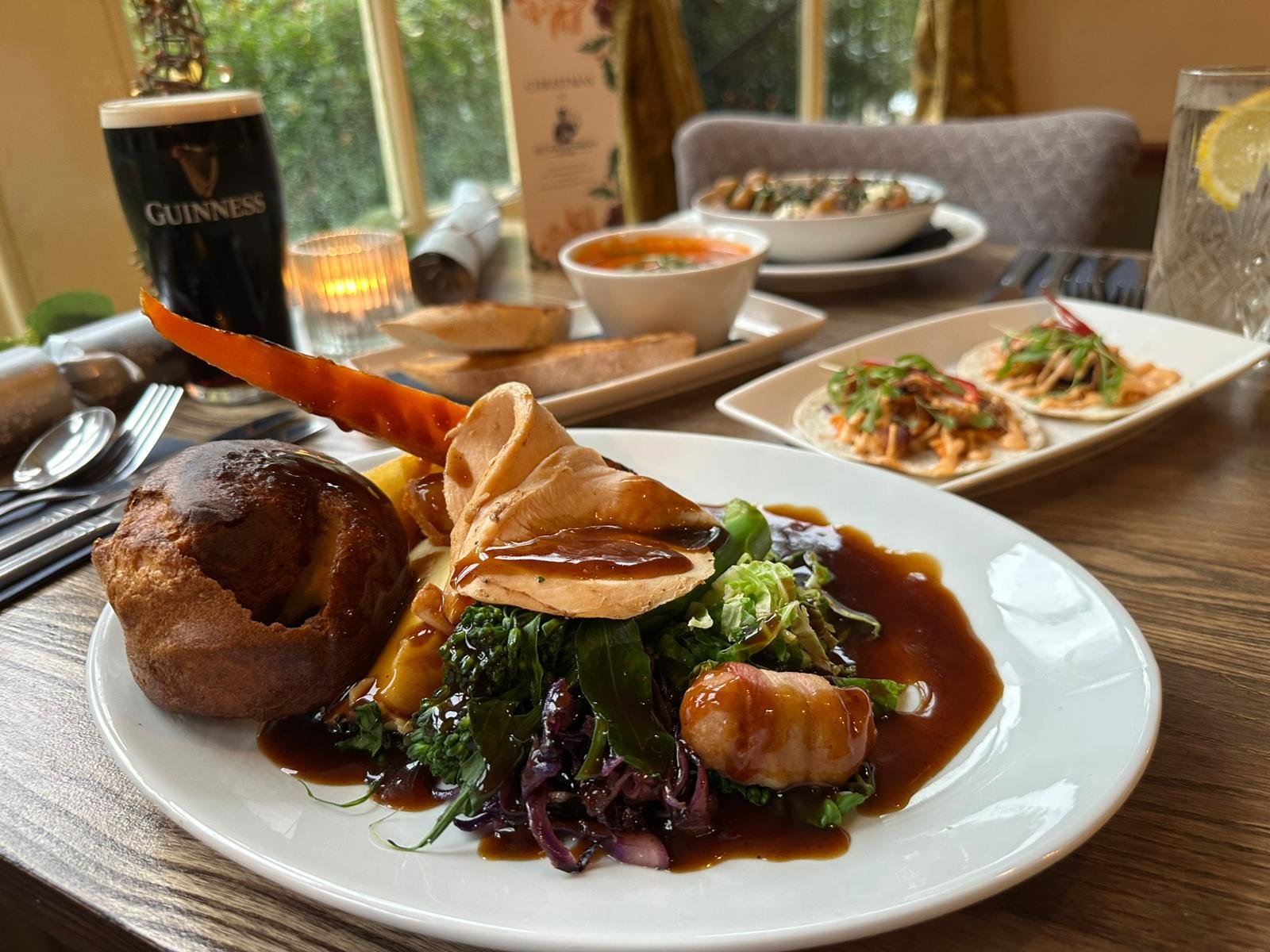 A plate of gourmet food with roasted chicken, stuffing, and mixed greens with gravy, served on a wooden table. In the background, there are plates of appetizers, glasses of water and soda, and a Guinness beer. The setting is a cozy restaurant with natural light from large windows.