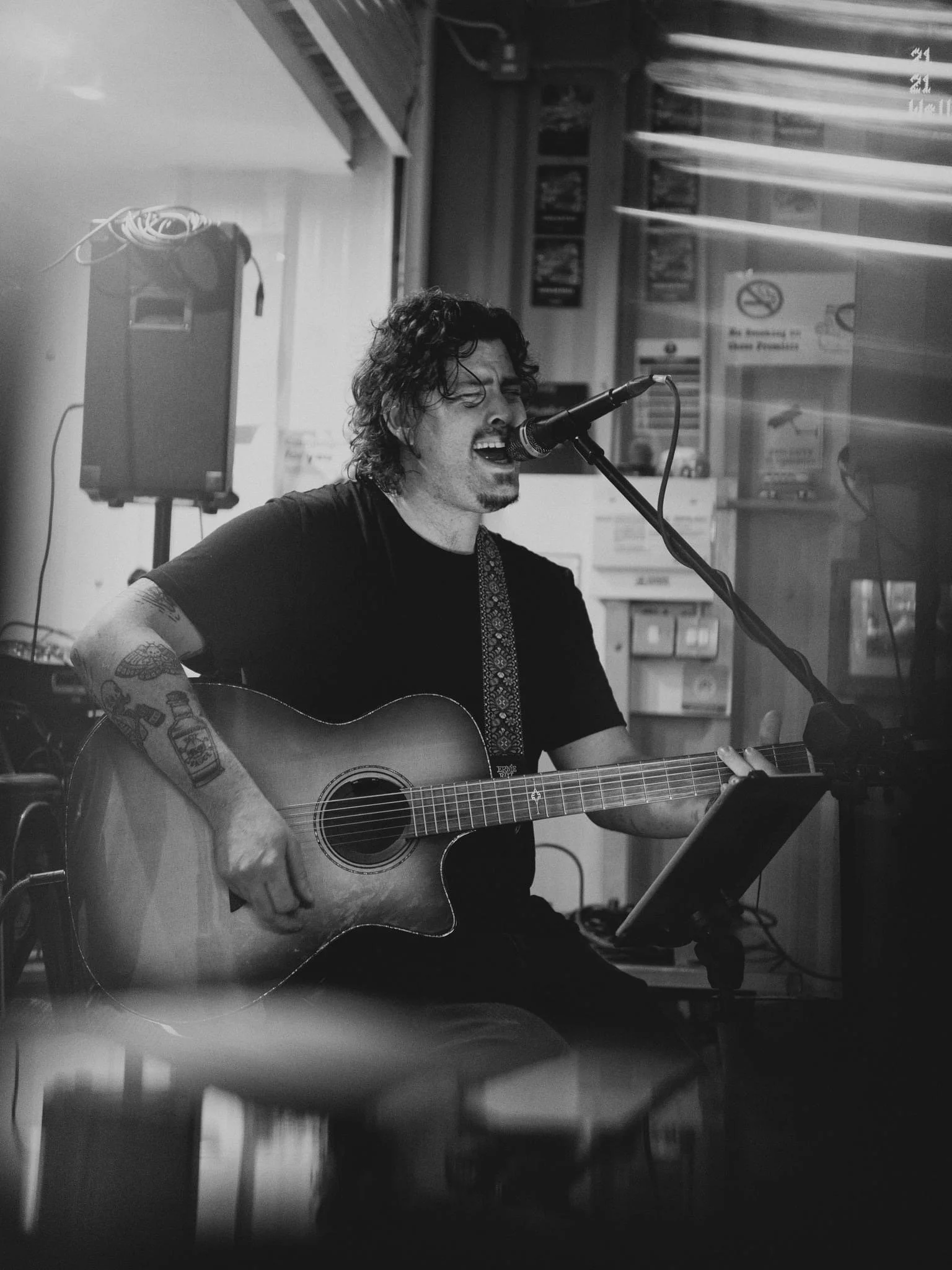 A man with curly hair and tattoos on his arm playing an acoustic guitar and singing into a microphone, in a cozy indoor setting.
