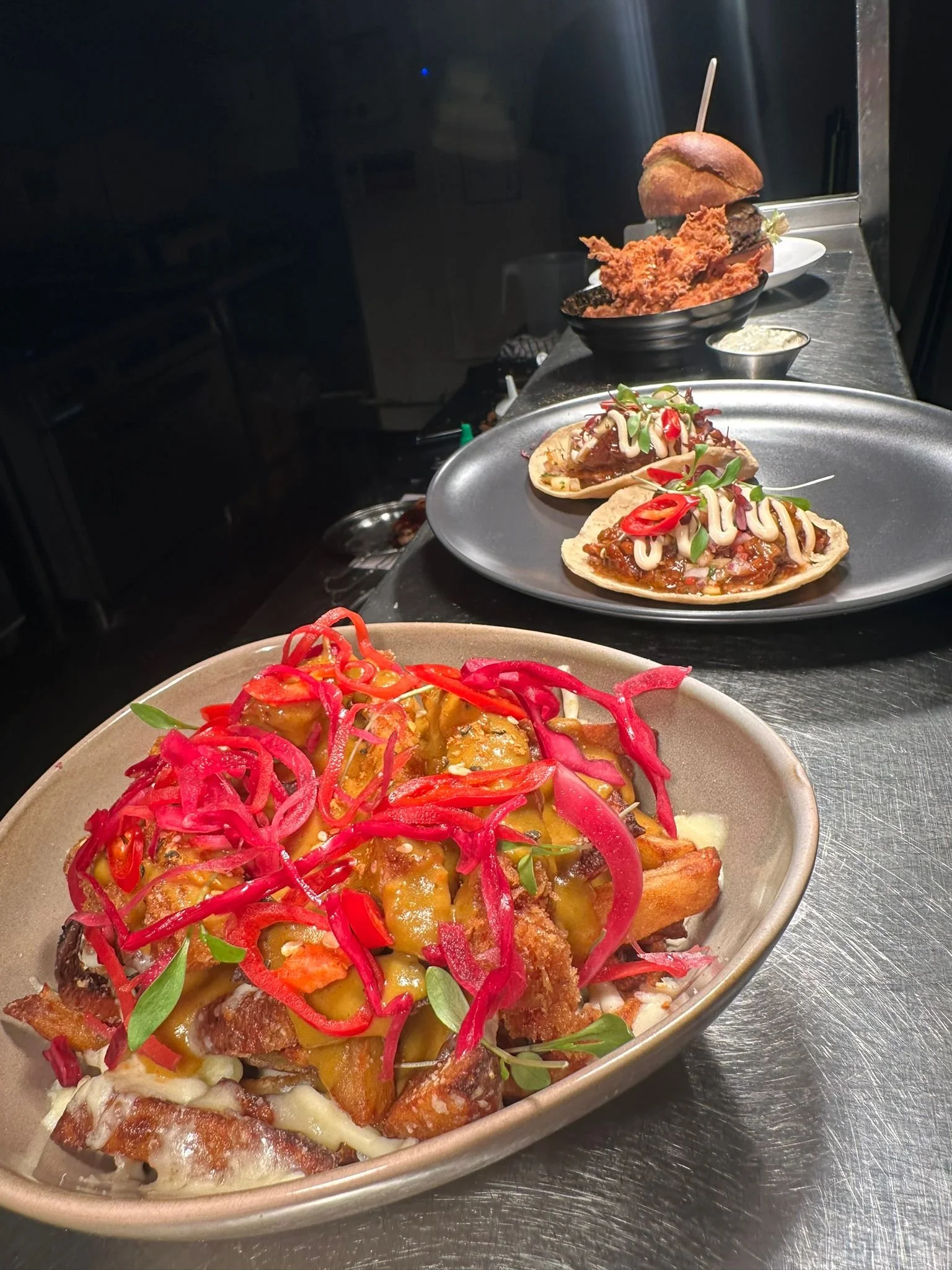 Dishes of loaded fries topped with cheese, pickled red onions, and herbs, two small tacos with meat and garnishes, a burger with fried chicken and a bun, and a bowl of fried chicken and a biscuit in the background.