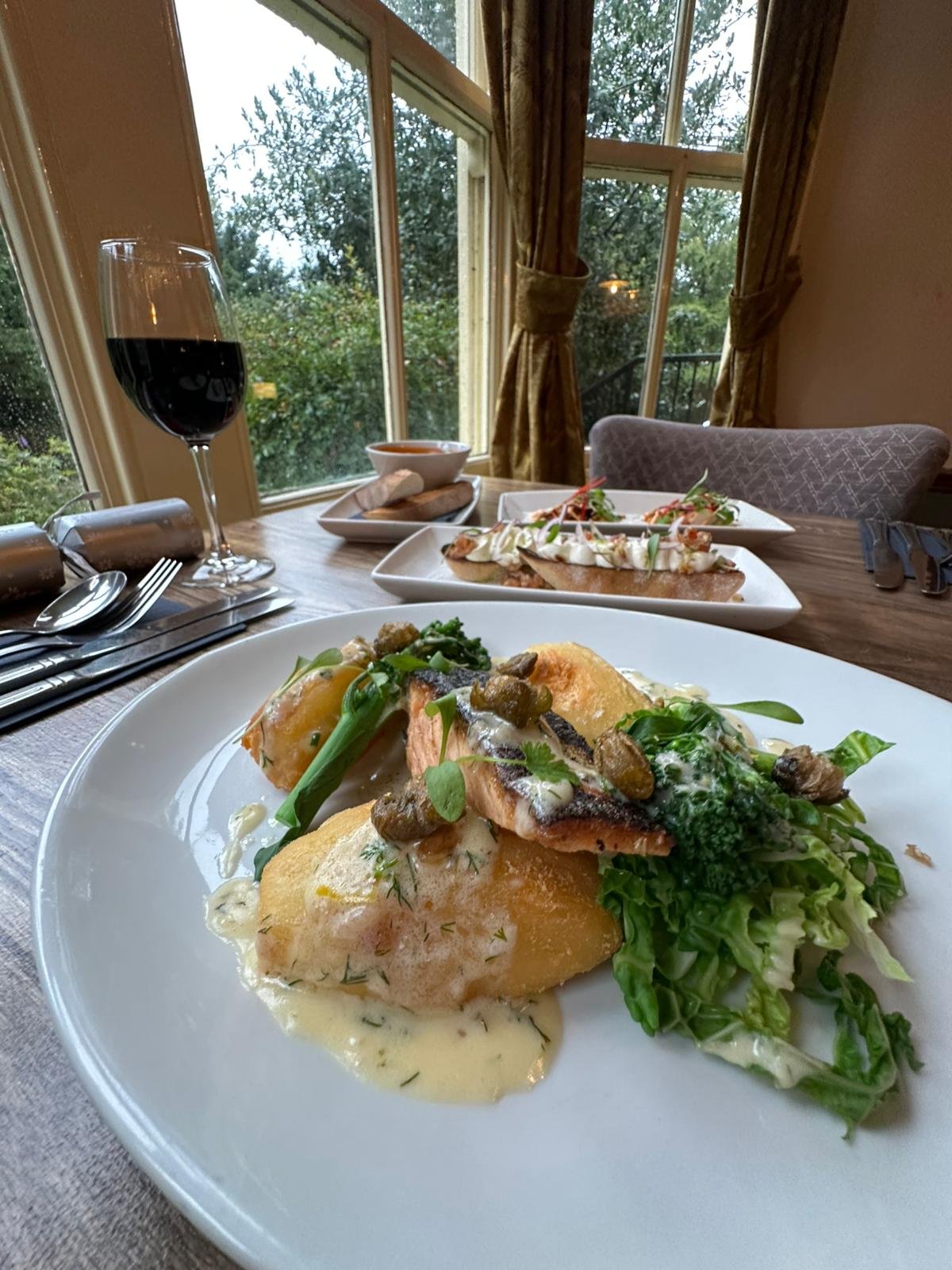 Plate with cooked fish, potatoes, and green vegetables at a dining table. In the background, there are additional plates of food, a glass of red wine, cutlery, and a window showing outdoor greenery.