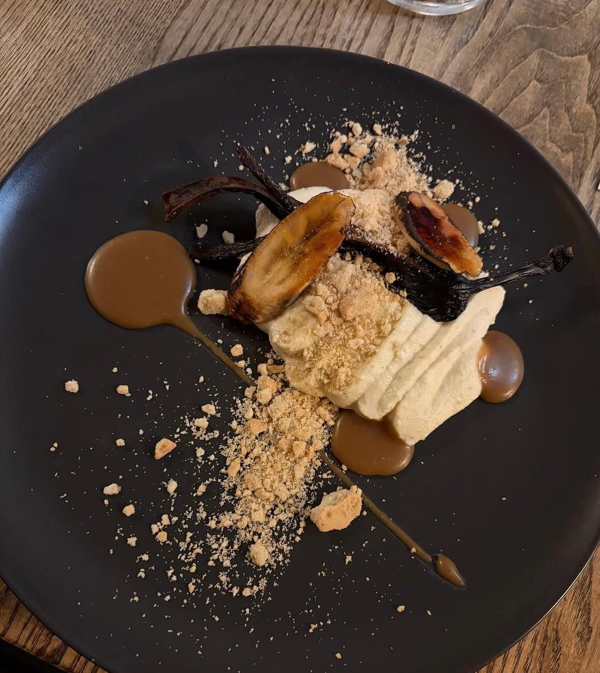A dessert plate with grilled banana slices, caramel sauce, whipped cream, crumbled cookies or biscuit crumbs, and chocolate sauce on a black plate.