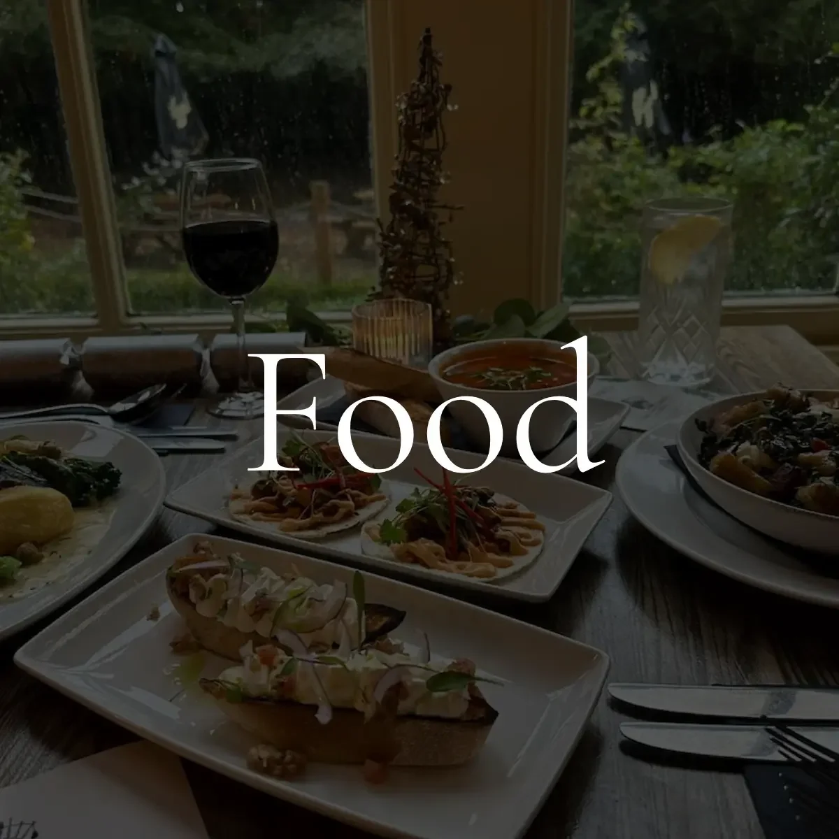 A table set with various dishes, including a glass of red wine, a bowl of soup, a glass of water with lemon, and plates of food, with a window showing greenery outside. The word "Food" is overlaid in white text.