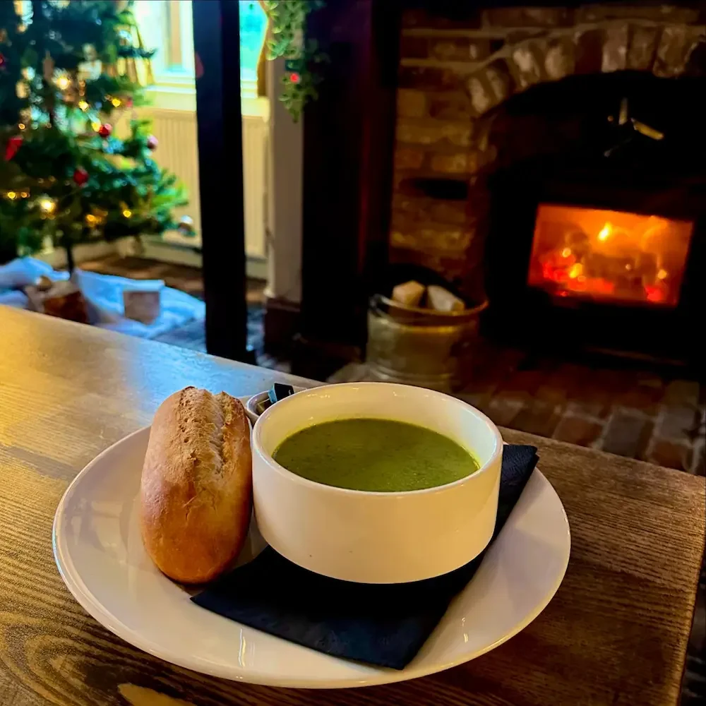 A bowl of green soup and a bread roll on a white plate with a black napkin, in a cozy room with a lit fireplace and a decorated Christmas tree in the background.