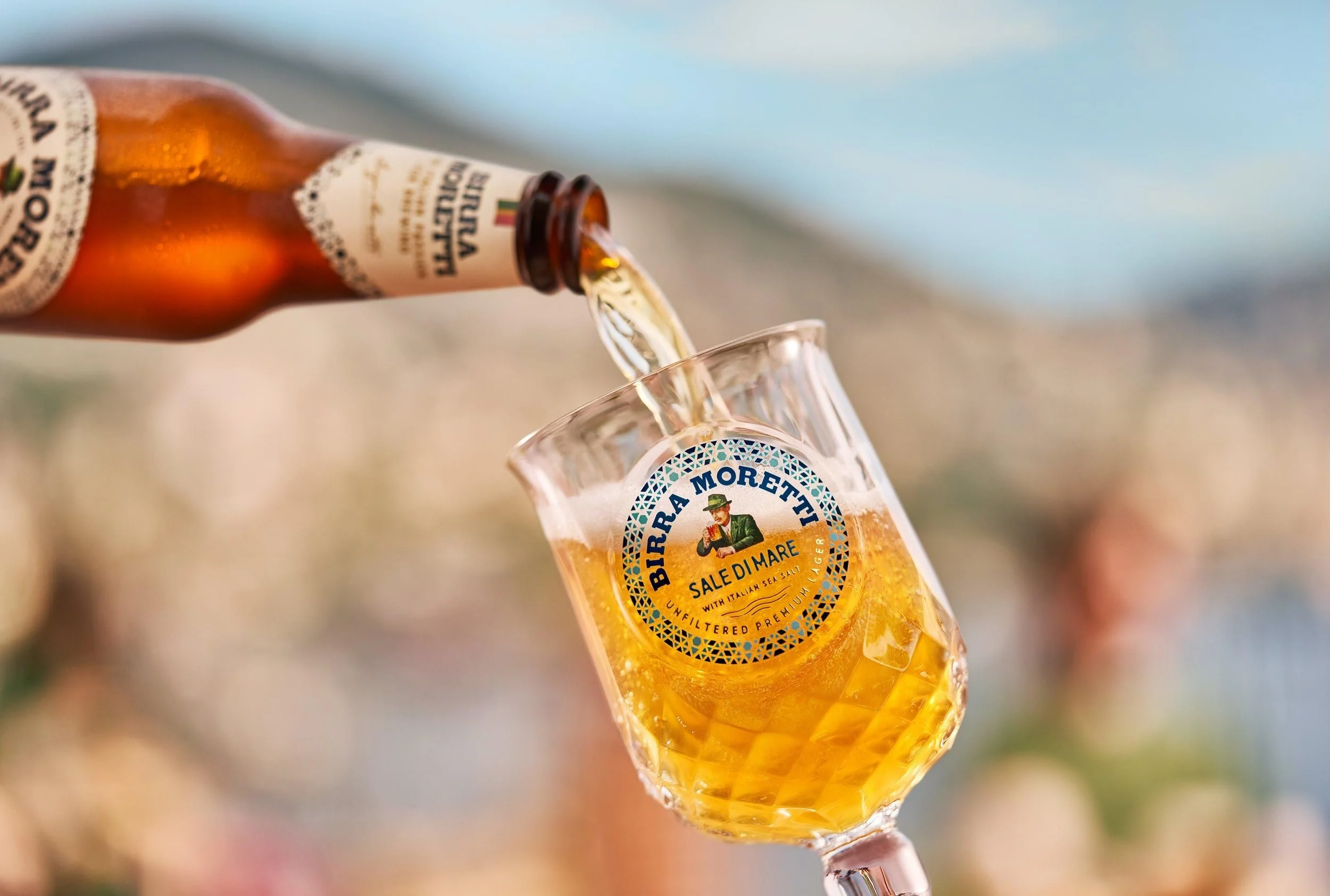 A bottle of Birra Moretti beer being poured into a glass with the label 'Birra Moretti Sale Di Mare' on it, with a blurred outdoor background.