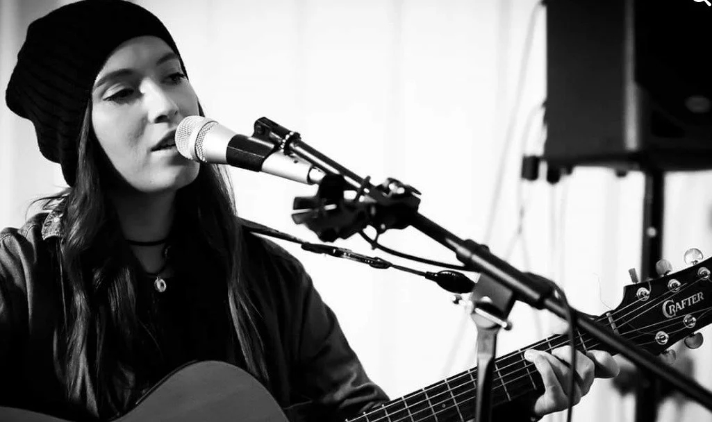 A young woman wearing a beanie hat and dark clothing playing an acoustic guitar and singing into a microphone.