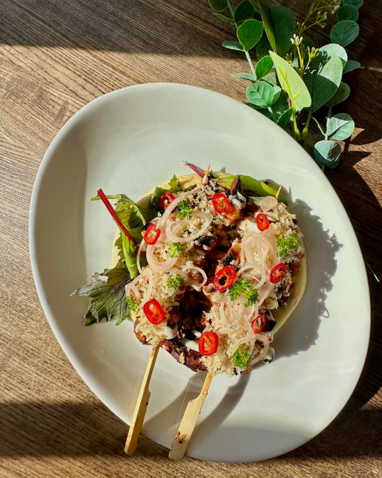 Two grilled skewers topped with sliced red chili, onions, herbs, and garnished with leaves on a white oval plate, with greenery in the background.