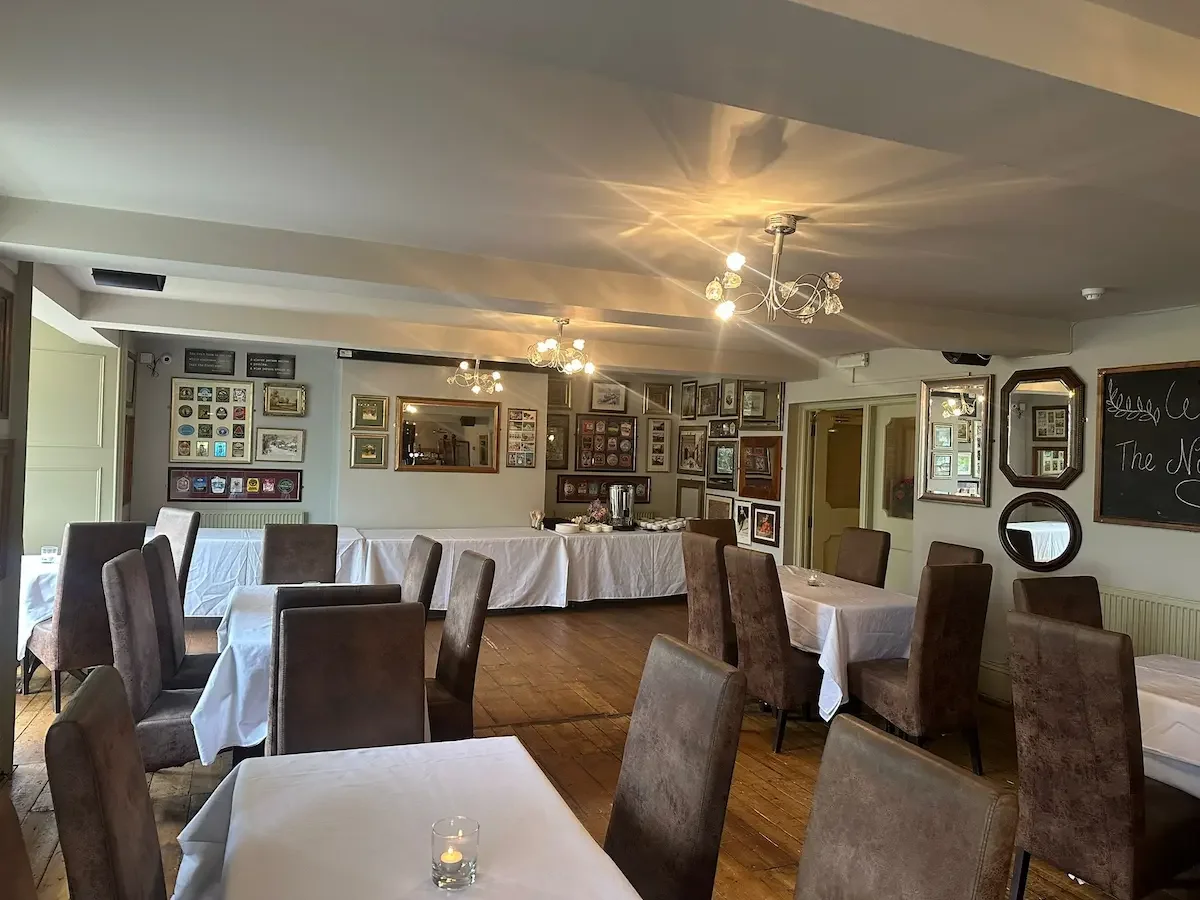 Interior of a cozy dining room with white tablecloth-covered tables, brown upholstered chairs, and a wall decorated with framed pictures and a chalkboard menu. Chandeliers hang from the ceiling, casting warm light.