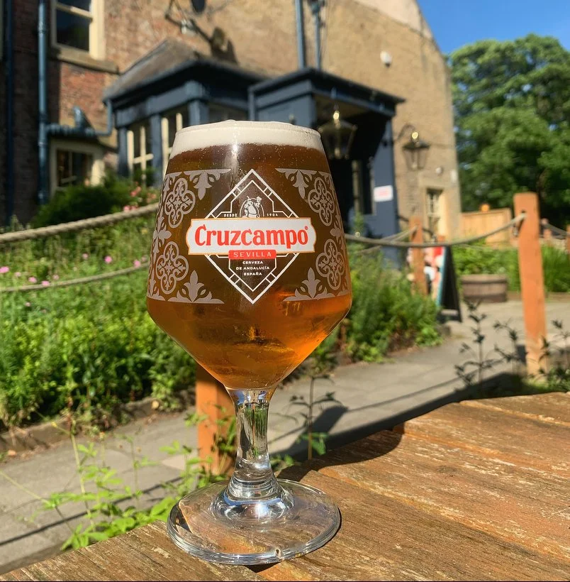 A glass of Cruzcampo beer with a foamy head, sitting outdoors on a wooden table, with a building and greenery in the background.
