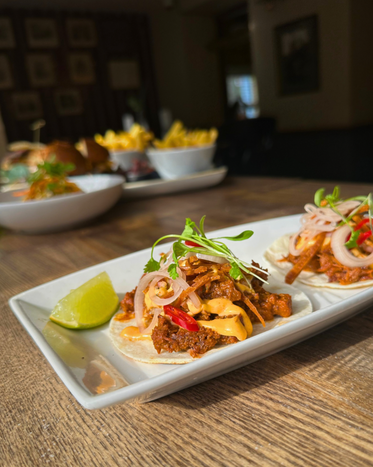 Close-up of two tacos with meat, sliced onions, sauce, and microgreens on a white rectangular plate, with a lime wedge on the side, on a wooden table.