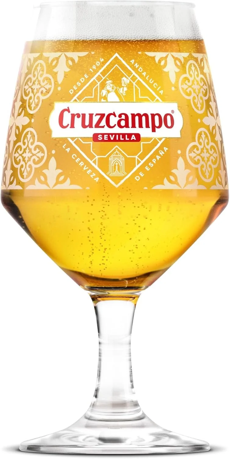 A glass of Cruzcampo beer with a decorative label that reads 'Sevilla' and features a white and gold pattern, with bubbles in the beer.
