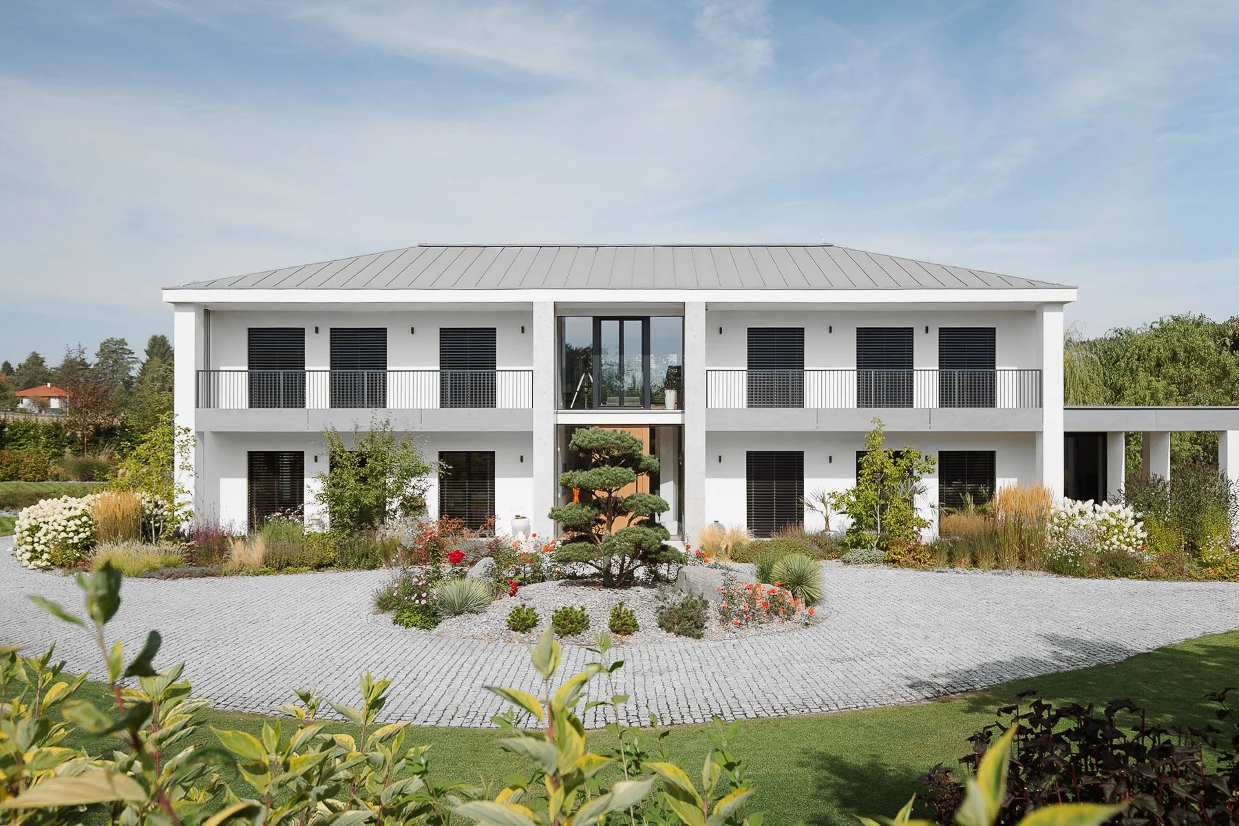 Exteriér moderní bílé dvoupodlažní vily s černými rámy oken, štěrkovou cestou a okrasnou zahradou.