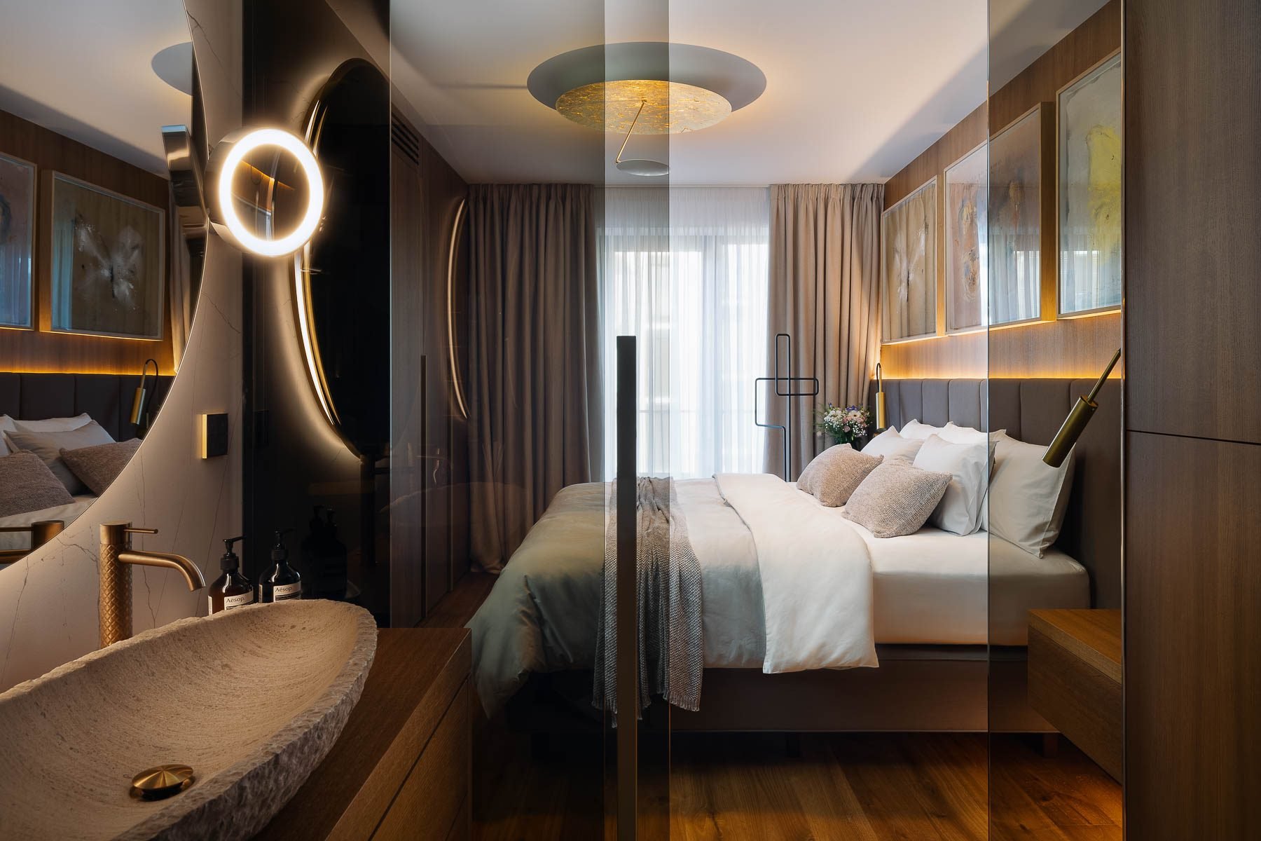 Moderní ložnice s designovým osvětlením a průhledem na kamenné umyvadlo přes skleněnou příčku