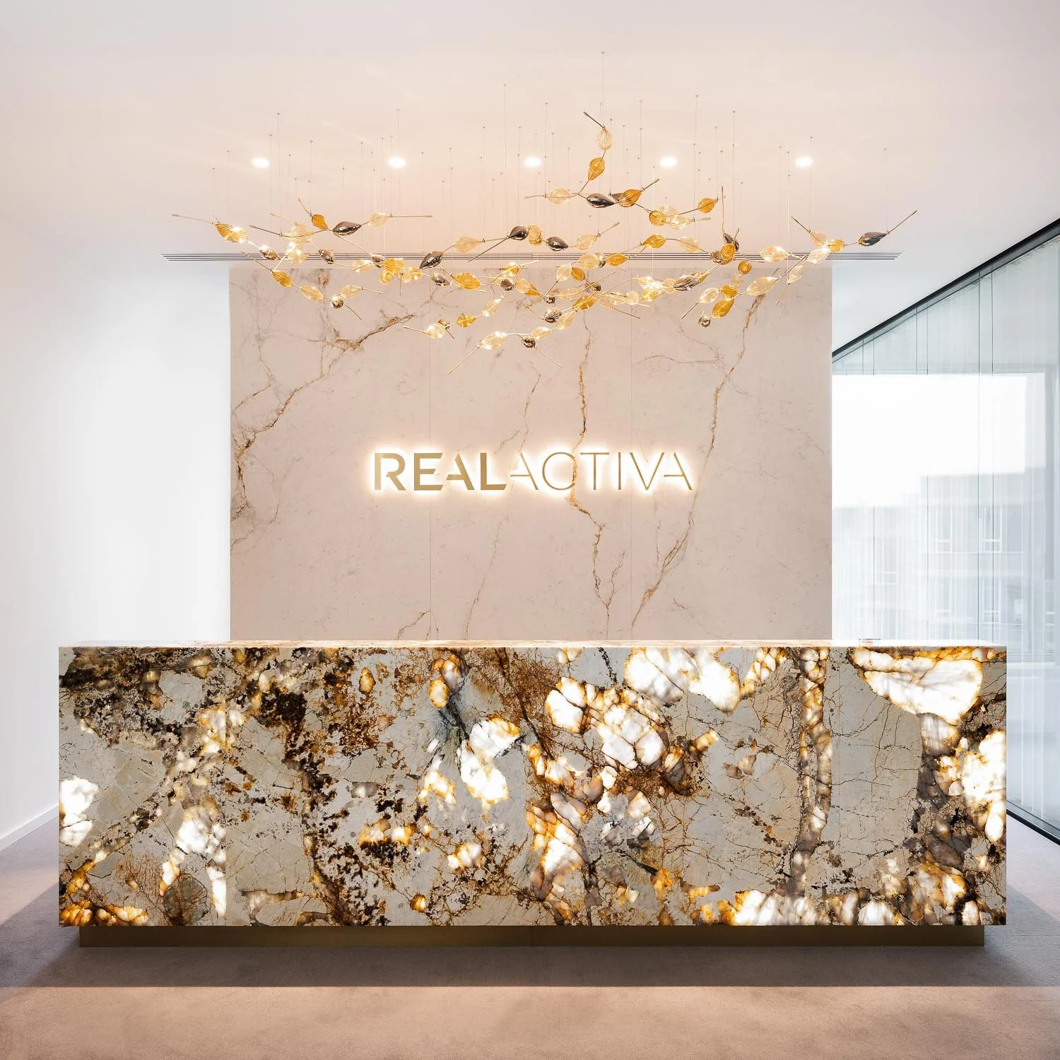 Světlá kancelářská recepce s logem „REAL ACTIVA“, podsvíceným kamenným pultem a dekorativní zlatou plastikou na stěně.