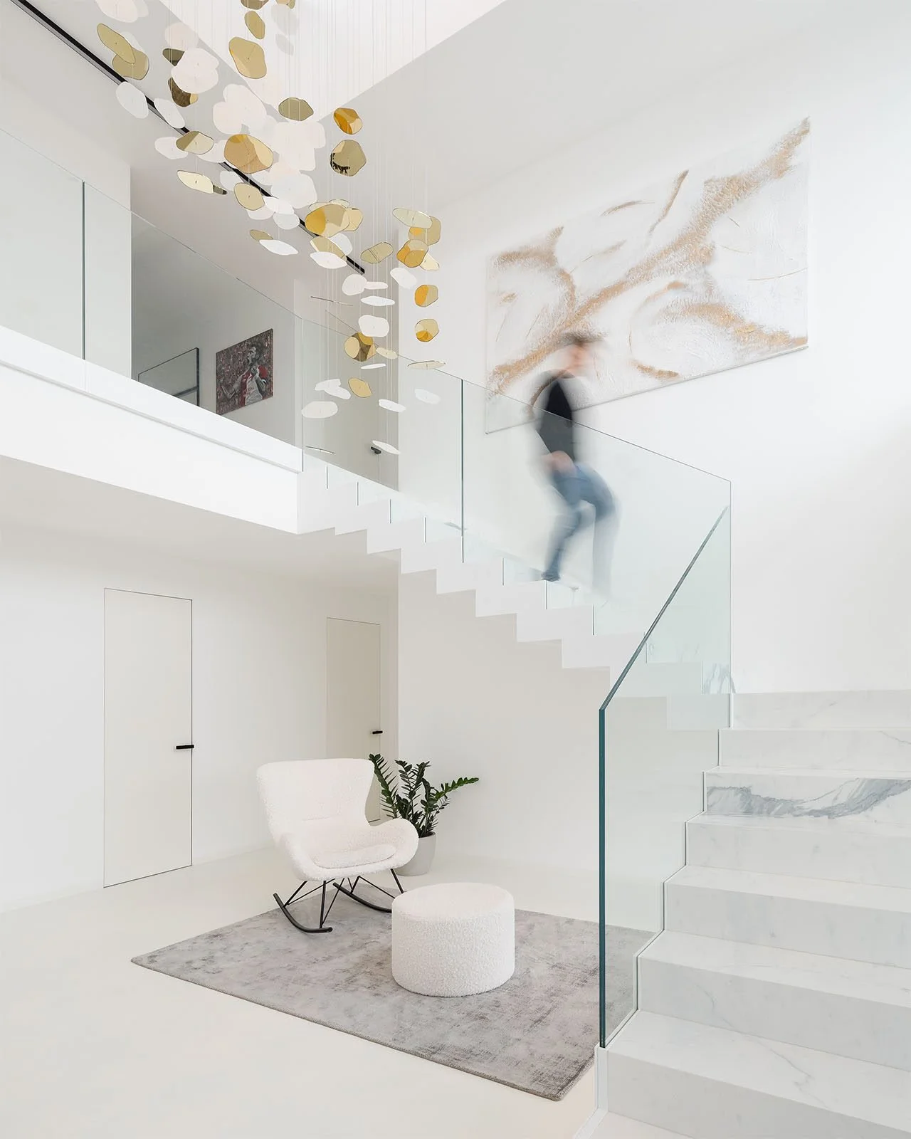 Světlý interiér s proskleným schodištěm, bílým designovým křeslem a moderním uměním na stěně.