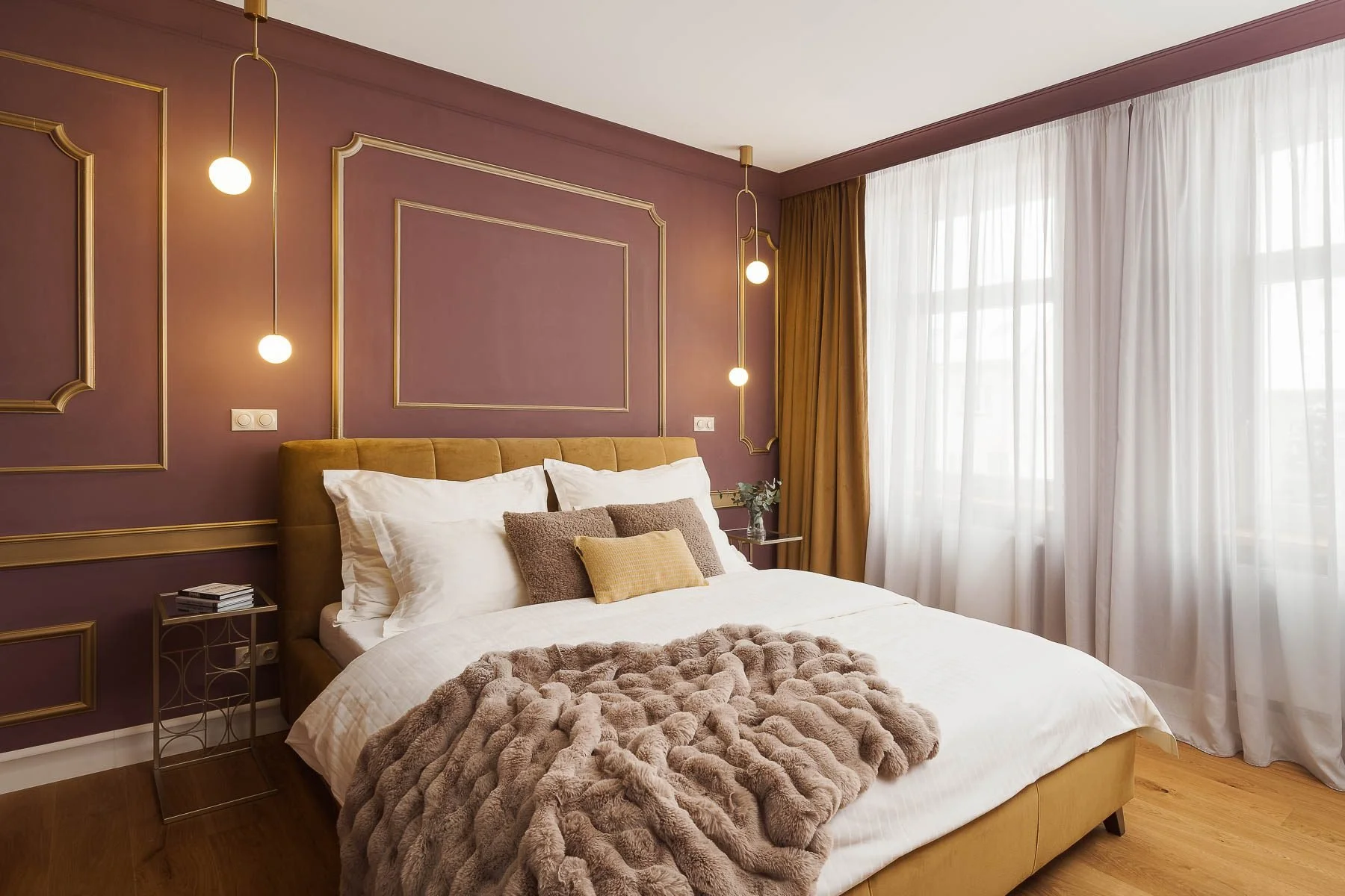 Interiér ložnice v kombinaci starorůžové a hořčicové barvy s ozdobným zlatým rámováním na stěně a huňatým přehozem.