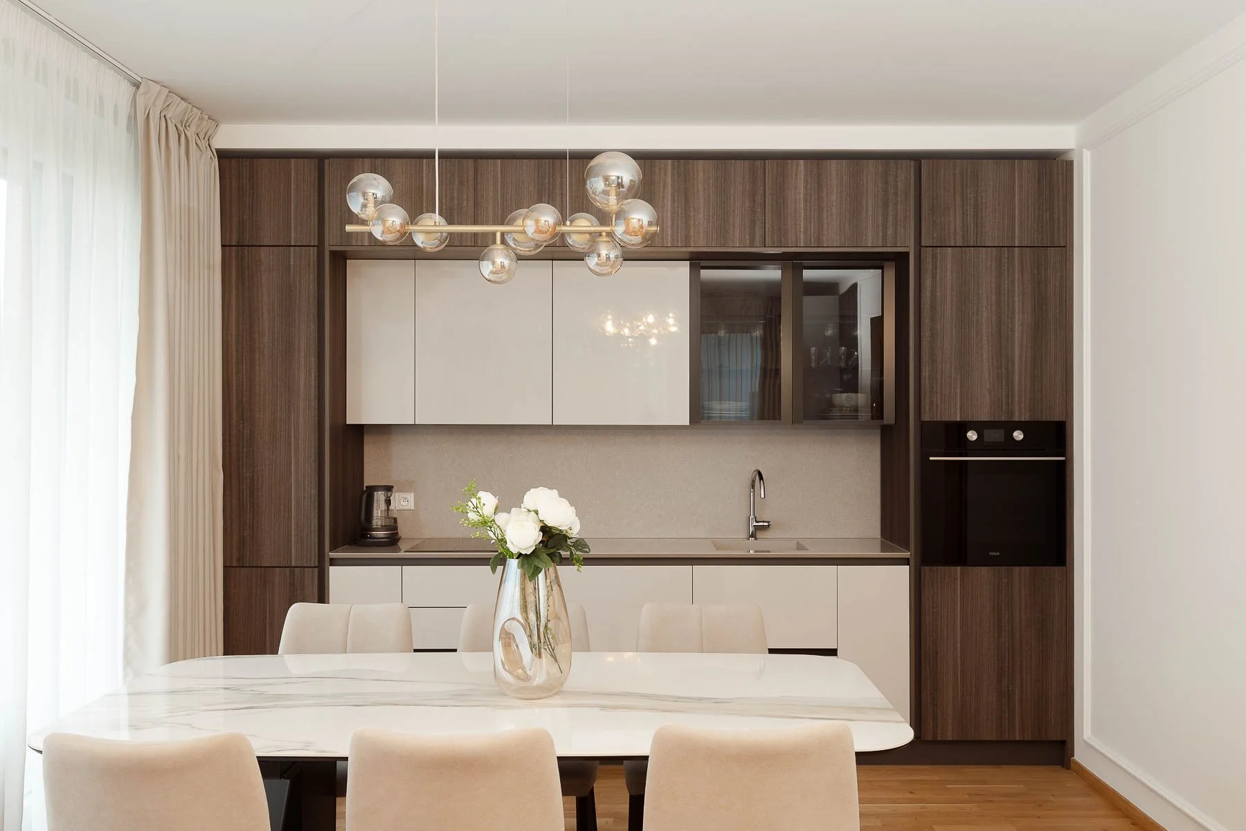 Jídelní stůl s mramorovou deskou, tmavý dřevěný dekor skříněk a designové kulaté svítidlo nad stolem.