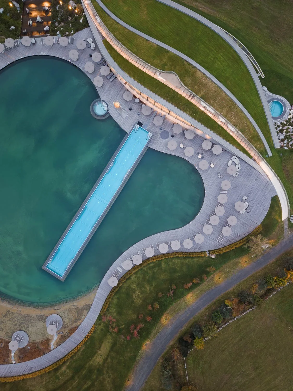 Letecká fotografie z dronu: kolmý pohled na designový infinity bazén, dřevěnou terasu a okolní zahradní architekturu resortu.