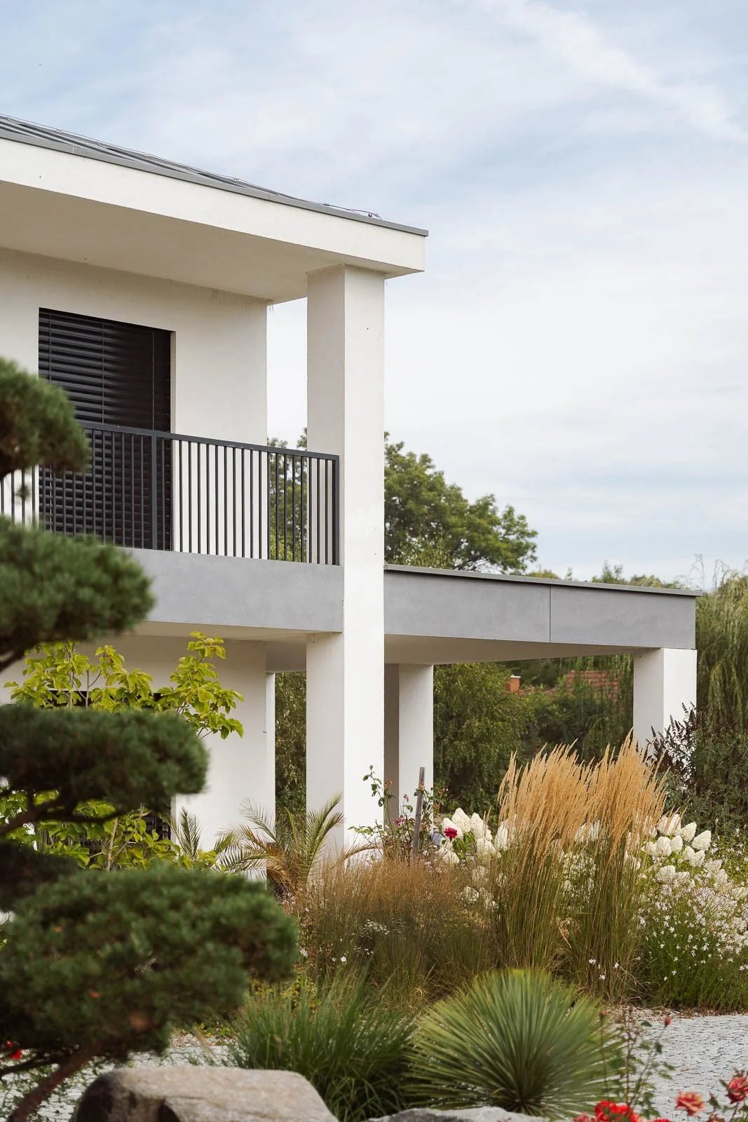 Pohled na terasu a balkon bílé vily s černým zábradlím, doplněný o vzrostlou zeleň a okrasné trávy.