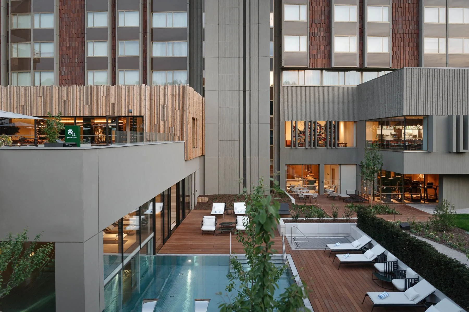 Pohled na venkovní relaxační zónu s modrým bazénem, dřevěnou palubou a bílými lehátky u moderní šedé budovy.