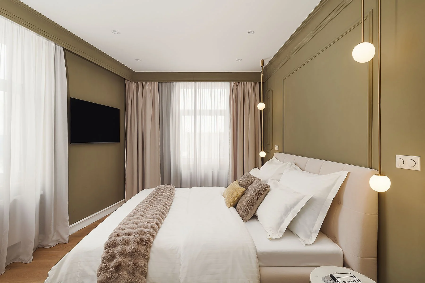 Moderní ložnice s olivově zelenou stěnou, čalouněným čelem postele a designovým osvětlením.