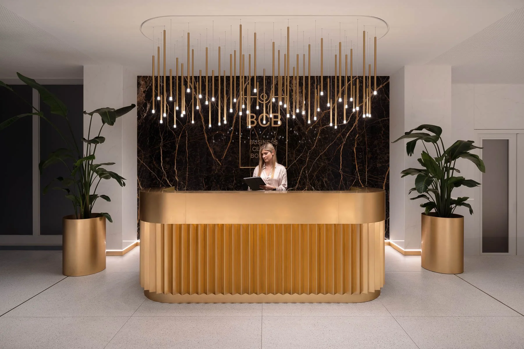 Moderní lobby s designovou zlatou recepcí, svislým lamelovým obkladem a efektní instalací svítidel.