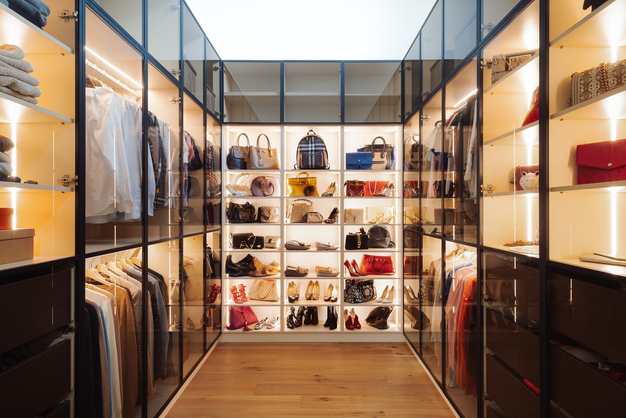 Prostorná šatna  s dřevěnou podlahou a prosklenými skříněmi, které mají podsvícené police pro vystavení obuvi a kabelek.