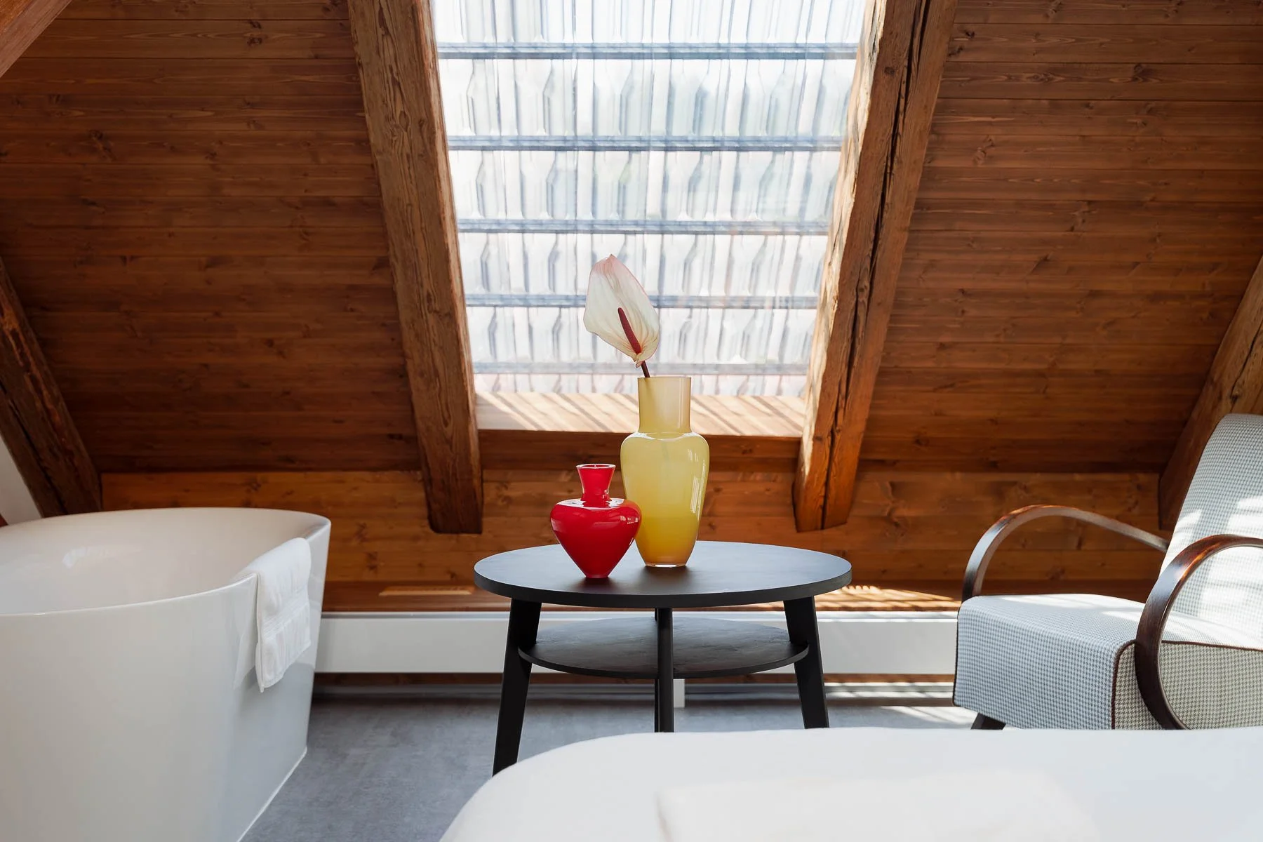 Designový interiér podkroví s dřevěnými trámy, střešním prosklením Nebesys, volně stojící vanou a uměleckými vázami Františka Jungvirta.