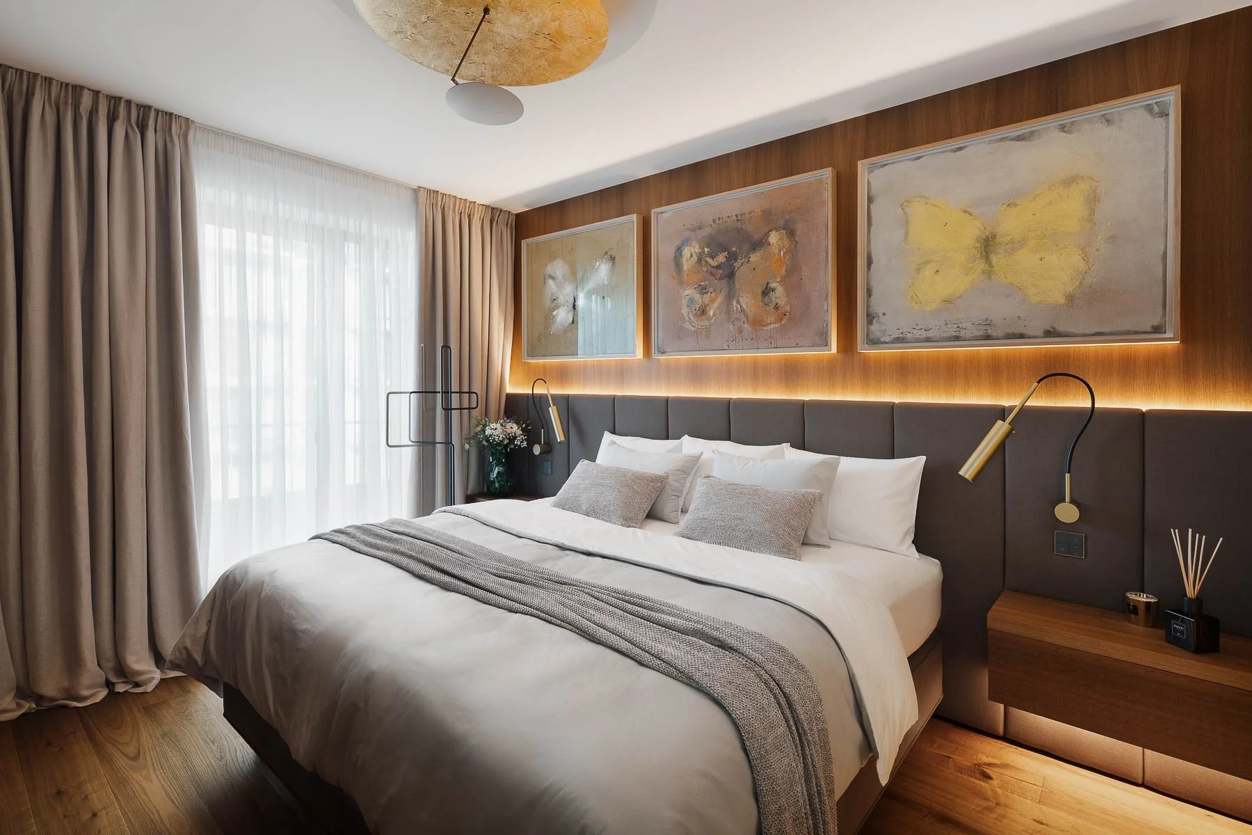 Interiér moderní designové ložnice s velkou postelí, abstraktními obrazy a dřevěnými prvky.