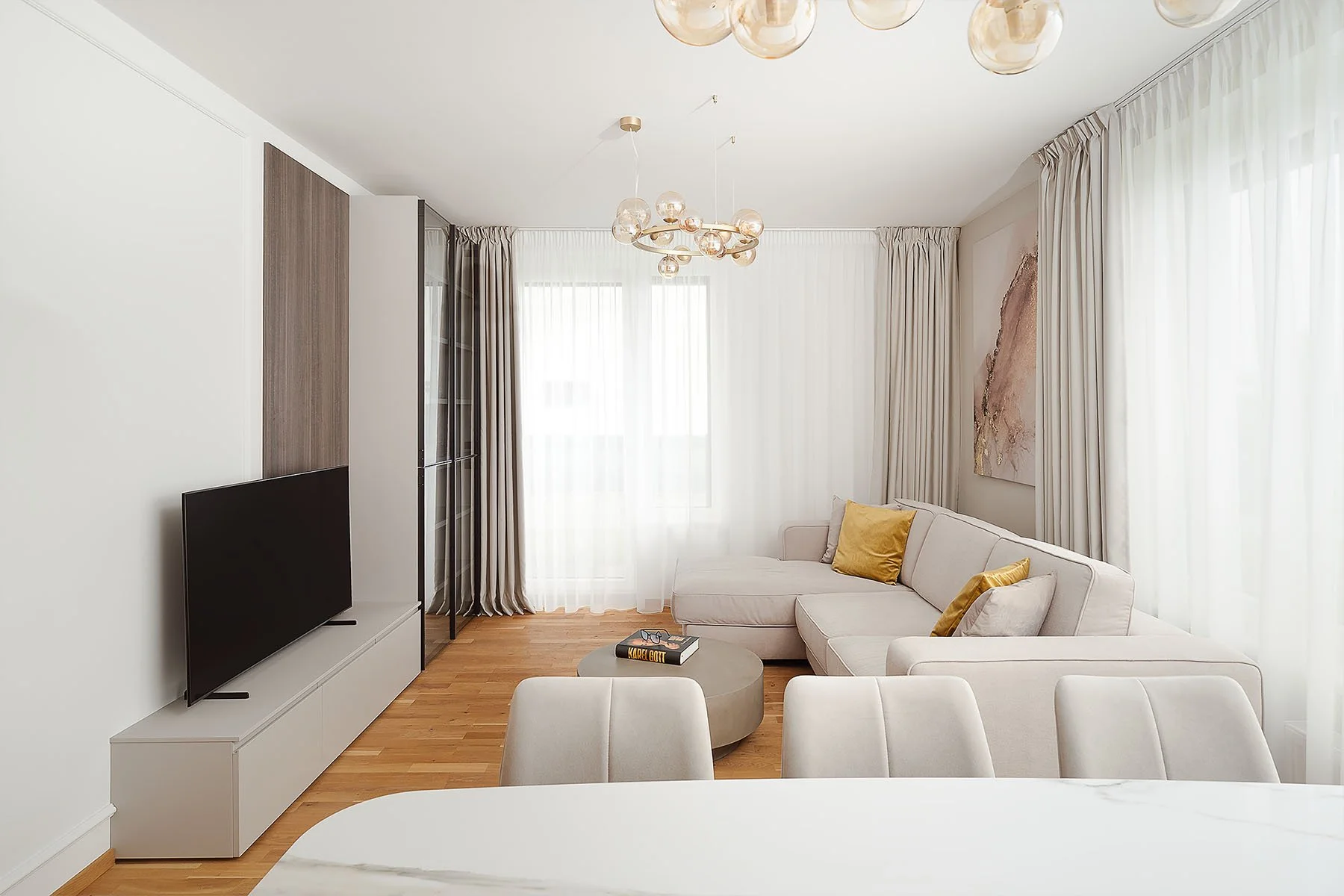 Prosvětlený interiér obývacího pokoje s velkými okny, světlou rohovou sedačkou a dřevěnou podlahou.