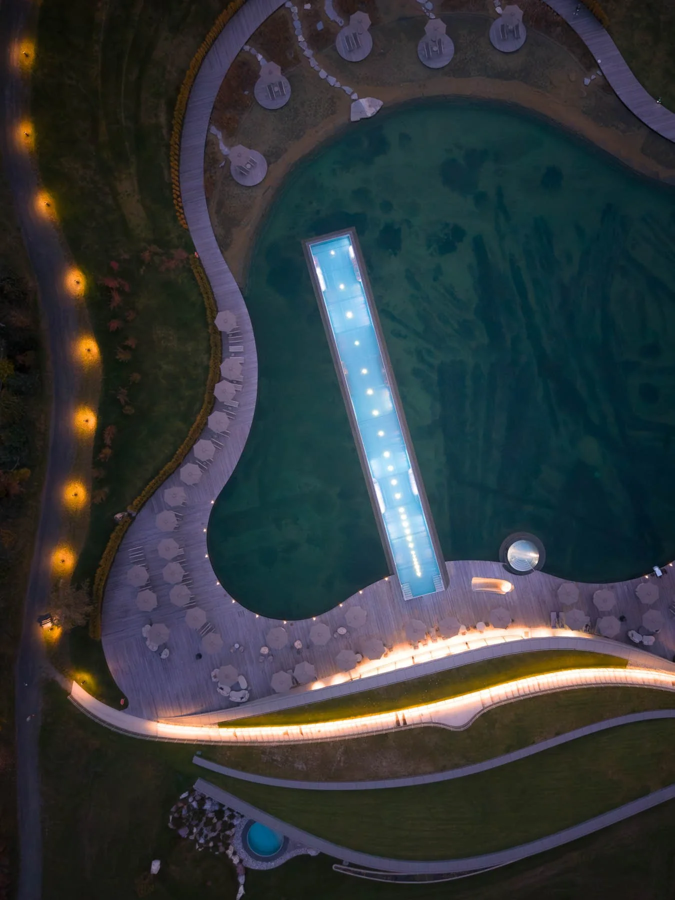 Letecká architektonická fotografie wellness Atmosphere by Krallerhof: osvětlený infinity bazén a přírodní biotop v moderním exteriéru při soumraku.